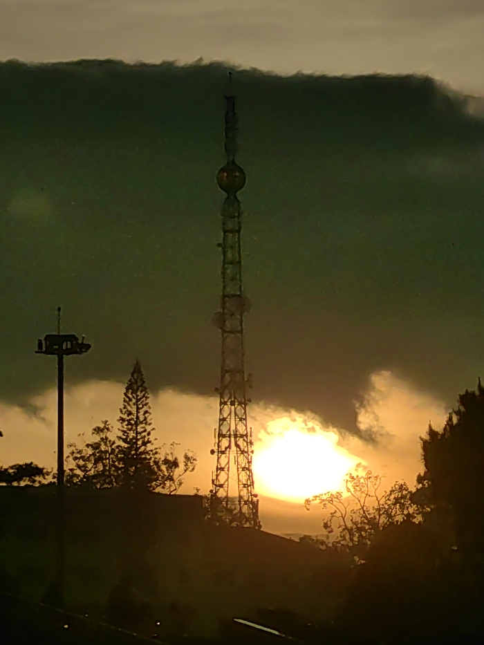 The image displays an elevated view of the Da Lat TV Tower (Tháp Truyền hình Đà Lạt) on Robin Hill in Da Lat, Vietnam, during twilight conditions, likely sunset. The primary subject is a tall, slender, lattice-structured communications tower, centrally positioned, featuring a distinctive spherical section near its summit, topped by a spire. This tower is silhouetted against a low, intensely bright sun, positioned in the mid-right of the frame and partially obscured by clouds.
To the left of the main tower, a shorter utility pole with a multi-directional fixture at its top is visible, also in silhouette. A prominent, slender pine tree stands between this pole and the main tower, and other indistinct, dark masses of foliage line the lower portion of the frame and the horizon. The sky exhibits heavy, dark green-grey cloud formations in the upper left, transitioning to vibrant orange and yellow hues around the sun. No human subjects or active interactions are depicted.