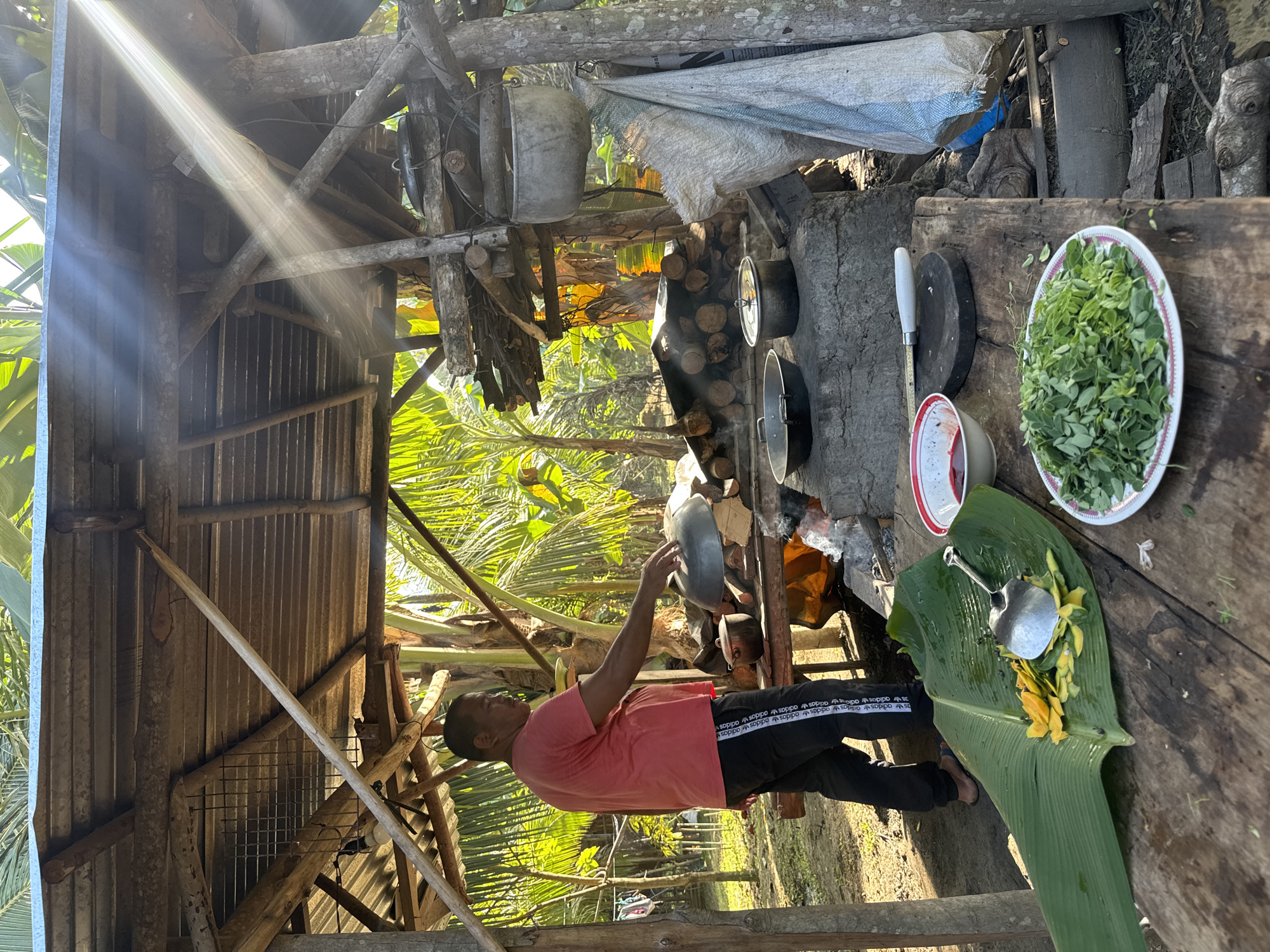 This image depicts an outdoor, rustic cooking area in Carmen, Philippines. A man, appearing middle-aged with dark hair, wears a coral-colored t-shirt and black track pants with white "Adidas" lettering. He stands on the left, barefoot on his right foot and wearing a blue slide sandal on his left, facing towards the right. His right arm is extended over a large, round metal pot or wok situated on a low, wood-fired stove structure, suggesting he is stirring or adjusting its contents.
The cooking area is sheltered by an open-air, unpainted wooden structure with a corrugated metal roof. The stove itself is constructed from concrete or stone, featuring multiple compartments for burning wood. Smoke is visible rising from the actively burning fire. Several metal cooking vessels, including pots and pans, are positioned on and around the stove, with additional pots hanging from the wooden framework. A stack of cut firewood is neatly arranged behind the stove.
In the foreground, a rough-hewn wooden table displays prepared ingredients and utensils. A large green banana leaf holds a pile of yellow fruit or vegetable peels and a rounded, silvery object, possibly a cleaned fish or dough, with a metal serving spoon resting on it. Adjacent to this, a white plate with a pink rim contains a substantial amount of finely chopped green leafy vegetables. Further right on the table, a white bowl with a red rim holds a dark red liquid and a green lime wedge. A small, dark circular cutting board and a knife with a white handle are also present.
The background reveals a lush tropical environment with dense green foliage, prominently featuring banana plants and palm trees. Strong, direct sunlight filters through the upper right of the scene, casting distinct rays and illuminating the vibrant greens. Various utilitarian objects, including large woven sacks and cloths, hang from the wooden support beams on the right side of the frame. In the far left background, clothes are visible drying on a line. The ground beneath the structure is exposed earth.