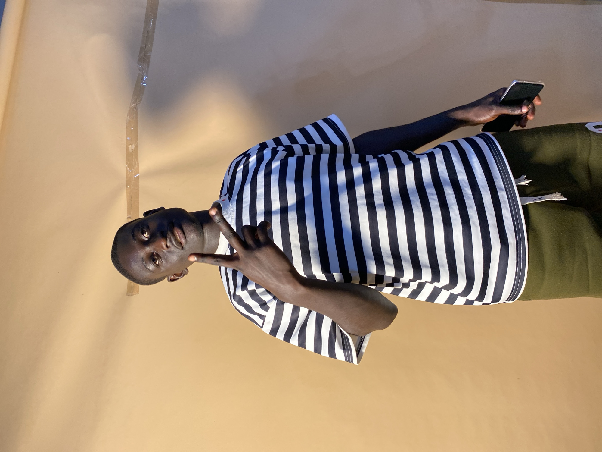 A person stands in front of a tan or beige backdrop, holding a black smartphone in their right hand and making a peace sign with their left. The person is wearing a black and white striped shirt and what appears to be olive-green pants or shorts. The scene includes a wall and lighting effects. The exact location in Bukuru, Nigeria, cannot be determined from the image alone.