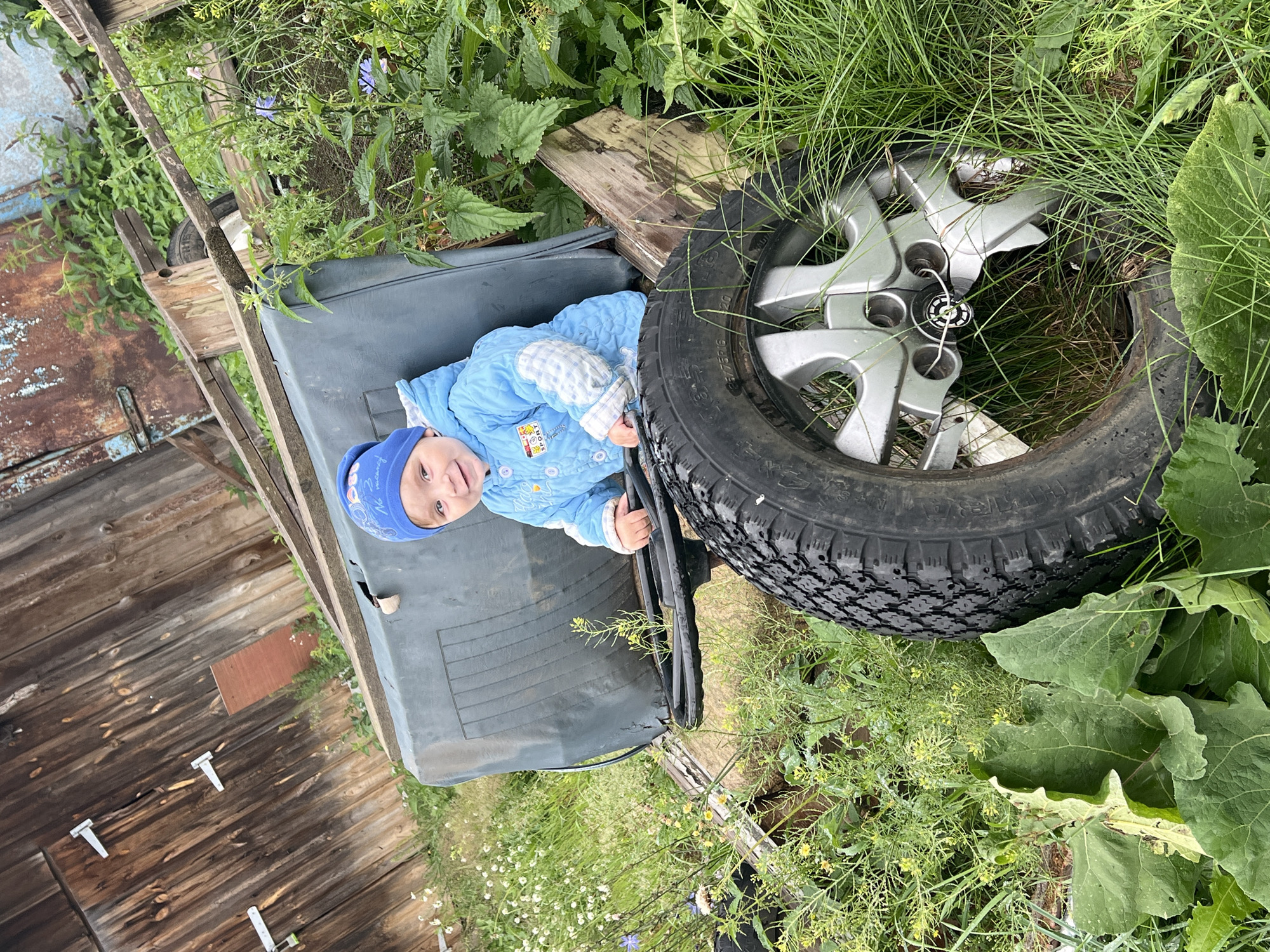 A young child, wearing a blue quilted jacket and a blue beanie, sits in what appears to be the driver's seat of an abandoned vehicle or piece of machinery, gripping a black steering wheel. The child is smiling. The vehicle's seat is a dark, weathered fabric, and a large, worn tire with a silver hubcap is positioned in the foreground. The setting is outdoors, surrounded by tall green grass, weeds, and some wildflowers. In the background, a weathered wooden structure, possibly a barn or shed, with visible hinges and planks, is visible, as is a rusted metal surface. The overall scene suggests a rural or derelict location, consistent with the outskirts of Panteleevka, Russia, where such discarded machinery and structures are common.