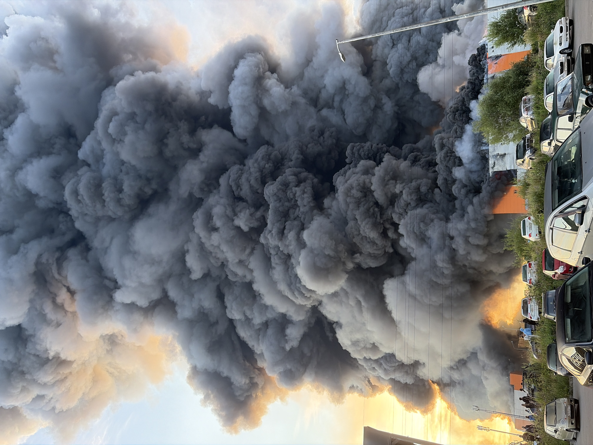 A massive column of dark grey smoke billows into a pale blue and orange sky from a large building fire in Nikolskoye, Russia. The smoke plume is dense and voluminous, obscuring much of the sky and creating a dramatic, apocalyptic scene. At the base of the smoke, flames are visible, licking up the side of an orange and white striped building. Several cars are parked in front of the building, some obscured by the smoke and foliage. A few people are standing near the cars, observing the fire. A street lamp stands tall to the right of the smoke column. The overall impression is one of an intense and uncontrolled inferno.