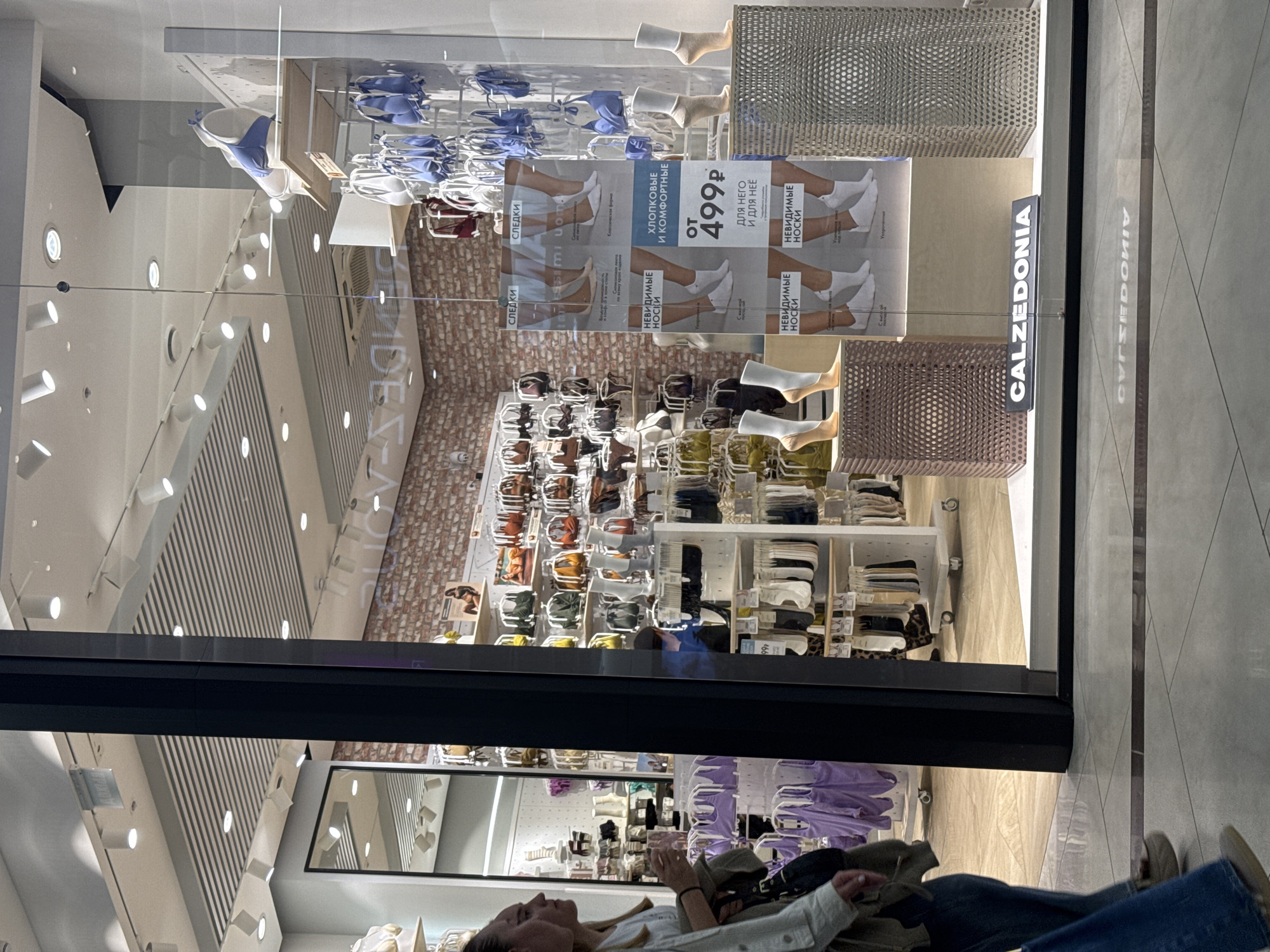 The image depicts a storefront of a Calzedonia store within an indoor shopping mall in Perm, Russia. The display window showcases lingerie and hosiery. In the upper section, blue and some maroon bras and underwear are neatly arranged on racks.
A large promotional poster dominates the central part of the display. It features various styles of white socks, referred to as "НЕВИДИМЫЕ НОСКИ" (Invisible Socks) and "СЛЕДКИ" (Footies). The text highlights them as "ХЛОПКОВЫЕ И КОМФОРТНЫЕ" (Cotton and Comfortable) and available "ДЛЯ НЕГО И ДЛЯ НЕЁ" (For Him and For Her), starting "OT 499P" (from 499 rubles). Several mannequin feet wearing these white socks are incorporated into the display. The background of the display includes a brick-patterned wall.
The lower section of the window contains shelves with folded items, likely other apparel or accessories, in neutral tones such as brown and olive green.
The reflective glass of the storefront clearly mirrors the interior of the shopping mall. The white ceiling with numerous spotlights and strip lights is visible in the reflection. A person is reflected sitting or leaning, wearing blue jeans and a light-colored top, looking towards the store. The tiled floor of the mall walkway is also visible in the reflection. The store's name, "CALZEDONIA," is displayed above the window and is also faintly reflected in the glass. The scene suggests a typical retail environment during shopping hours.