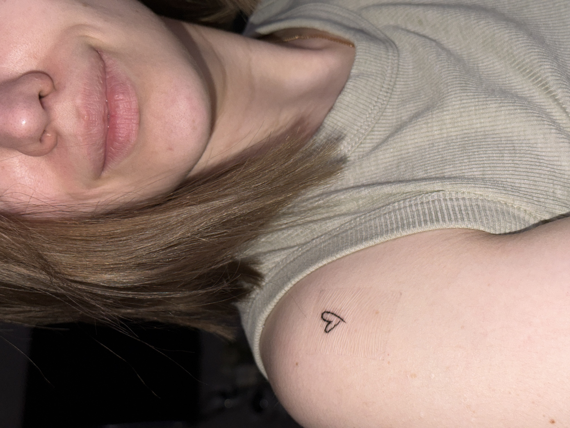 The image shows a person from the chest up, slightly tilted to the left. The person has fair skin, brown hair, and pink lips. A small black outline of a heart is tattooed on their upper arm. They are wearing a ribbed, light green top. The background is dark and out of focus. There is no indication this image was taken in the city of Reshetnikovo, Russia.