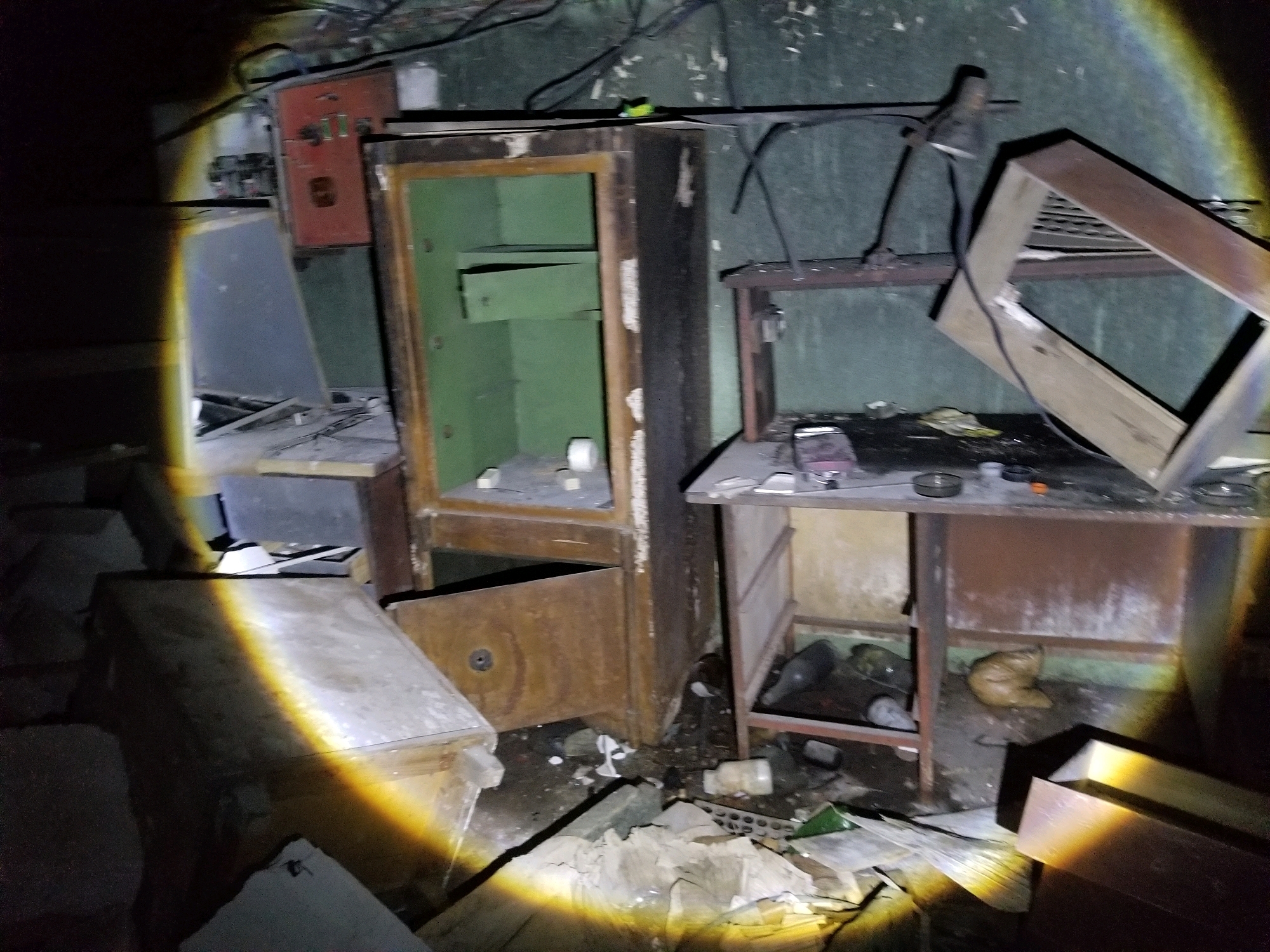 The image depicts an abandoned room with a cluttered environment. In the foreground, there is a large, open wooden chest or cabinet. To the left and right of the chest are various discarded items, including what appear to be pieces of wood, paper, and debris. A strong yellow light beam illuminates parts of the scene from the bottom left.
In the mid-ground, there is a large, upright metal safe with its door ajar, revealing a green interior with shelves. Adjacent to the safe, to the right, is a wooden desk or workbench. On this desk, several objects are visible, including what looks like a lamp with a flexible arm and a metal frame structure resting on it. There are also some containers and papers on the desk.
Further in the background, a red electrical panel is mounted on the wall. The walls and ceiling appear to be in disrepair, with exposed wires and a dark, grimy texture. The overall impression is one of neglect and disarray in an indoor, possibly industrial or storage space. There are no people visible in the image.