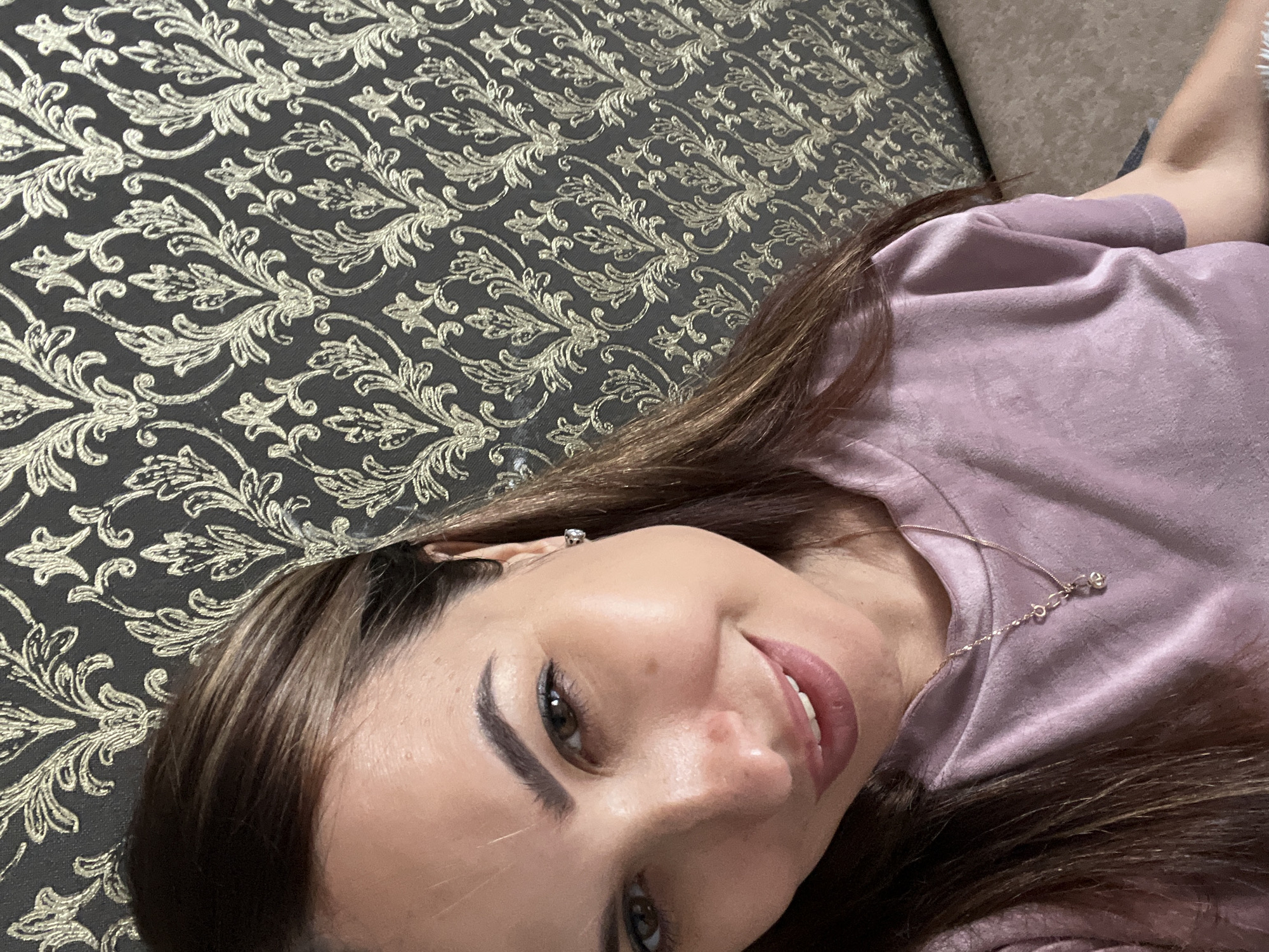 A close-up selfie shows a woman with long brown hair and brown eyes, smiling and looking at the camera. She is wearing a light purple, short-sleeved shirt and a delicate necklace. The background features a dark wallpaper with a repeating gold damask pattern. The lighting appears to be indoor.