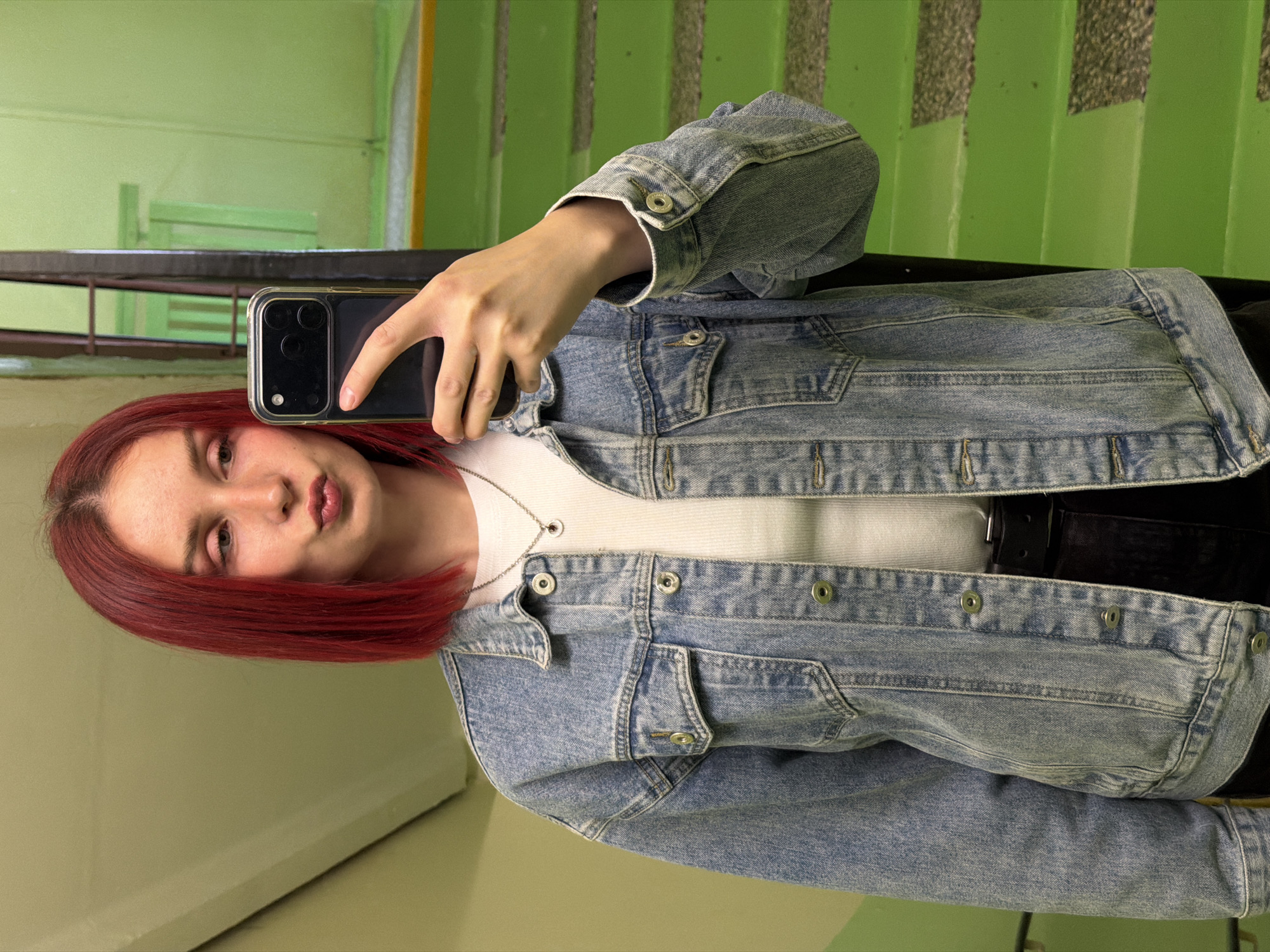 A young woman with red hair takes a selfie on a staircase in Chaikovsky, Russia. She wears a blue denim jacket over a white top and a silver necklace. She is holding a smartphone in a clear case, likely capturing her reflection in a mirror on the wall to the left. The background shows green-painted walls and stairs. The staircase is utilitarian, with a dark metal railing.