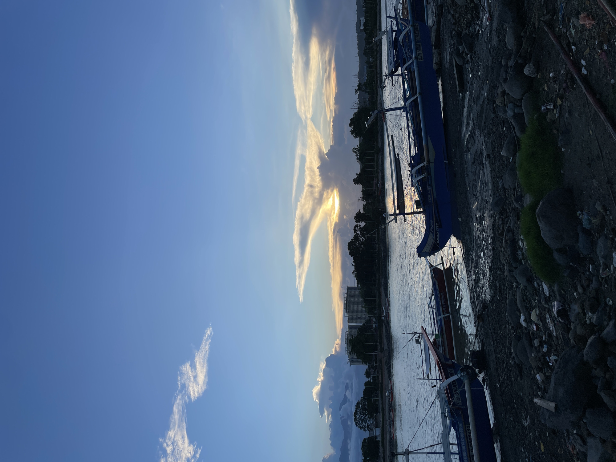 This is a medium-shot, eye-level photograph taken during twilight, showing a waterfront scene in Davao City, Philippines. The sky is a gradient of blue, transitioning to warm oranges and yellows near the horizon, with scattered clouds catching the last rays of the sun. In the distance, across a body of water, a line of trees borders the shoreline, with several buildings, including two prominent high-rise structures, visible behind them.
In the foreground, the muddy shore is littered with rocks, debris, and some sparse green vegetation. Several wooden outrigger boats are moored along the water's edge. The largest boat, painted blue and white, is prominent in the right half of the frame, its outriggers extending outwards. Another boat, red and white, is partially visible in the water to the left, and a third blue boat is closest to the viewer, its bow nudging the shore. The water reflects the soft light of the sky. No people are visible in the image. The overall impression is one of a tranquil, coastal urban environment at the close of day.