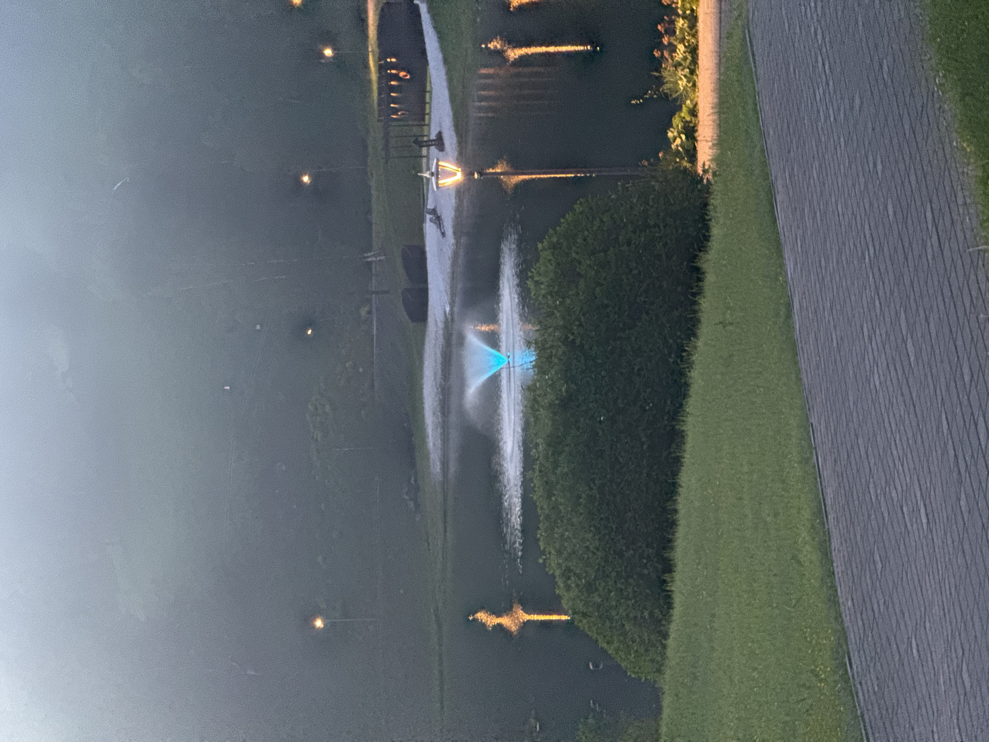 This image depicts a serene outdoor scene, likely in the city of Urlu-Aspak, Russia, characterized by a pond and a surrounding park-like environment. A lit fountain, emitting a vibrant blue light, is the central focal point in the pond. The water reflects the warm glow of several ornate street lamps positioned around the pond and in the background. Dense, dark green bushes and manicured green grass create a natural border around the water. A paved pathway, made of grey interlocking stones, occupies the foreground of the image. The background is shrouded in a soft, atmospheric mist, obscuring details of distant trees and structures, and suggesting either early morning fog or evening twilight. The overall impression is one of peaceful, illuminated natural beauty.