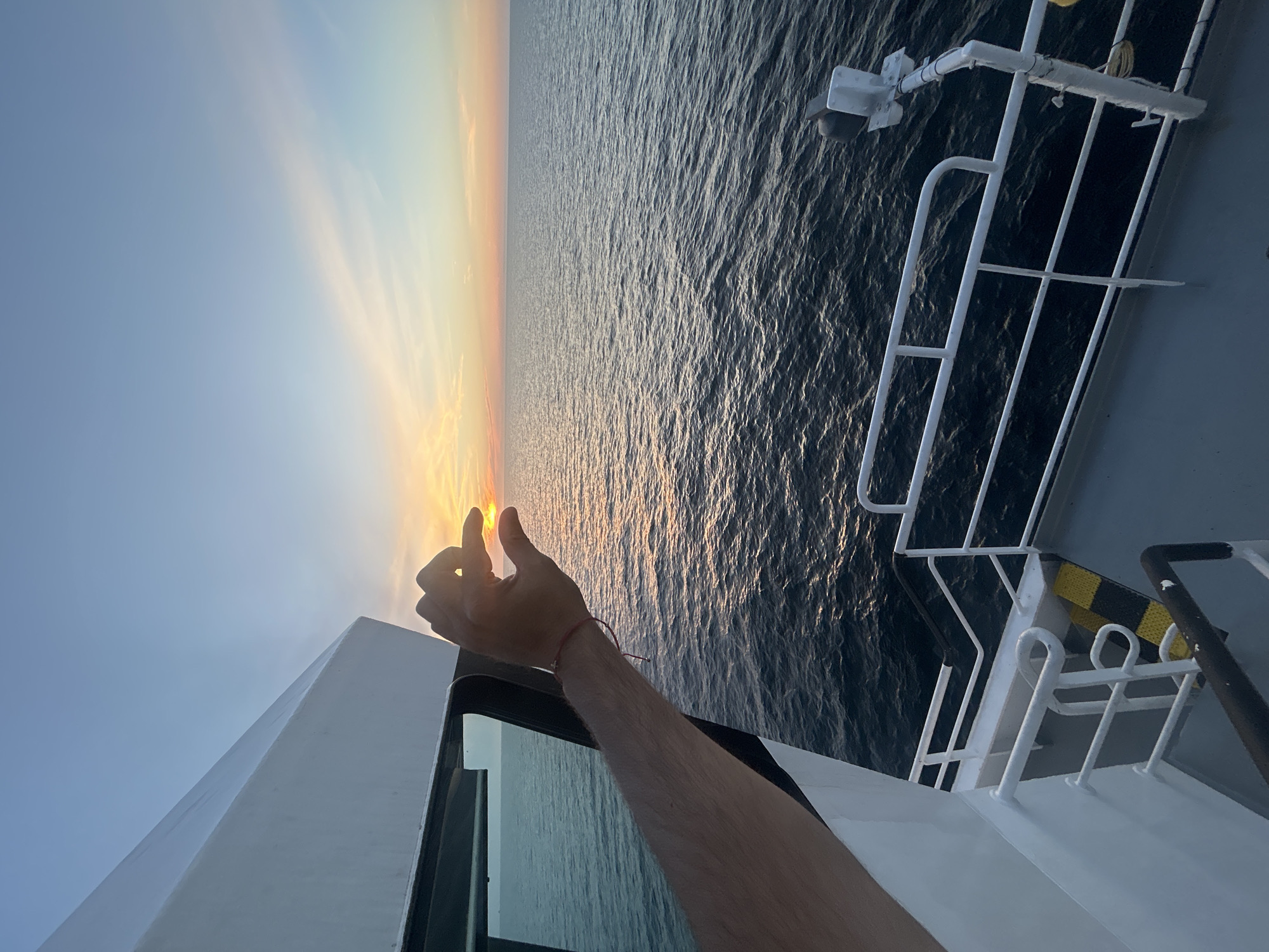 An arm with a red bracelet is extended from a ship's deck towards a sunset over the ocean. The sun is a small orange disc framed by the thumb and forefinger of a hand. The sky transitions from a clear light blue near the top of the frame to soft orange and pink hues near the horizon. The dark, rippling water of the ocean fills the lower half of the image, reflecting the sky's colors. The edge of a white ship structure, likely part of a cabin or bridge, is visible on the left, with a dark window. A white railing and part of a grey deck are also visible in the foreground. The image captures a moment of observing and interacting with the natural beauty of the sunset from the vantage point of a vessel at sea.