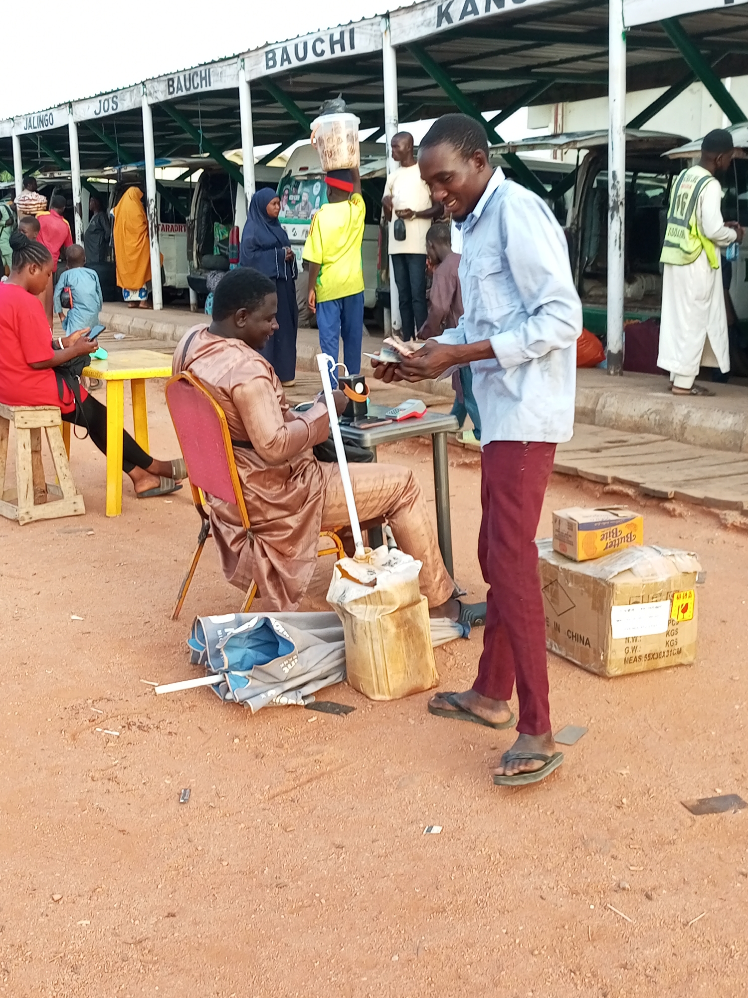 The image depicts a street scene, likely a bus park, in Gombe, Nigeria. In the foreground, a man in a light blue shirt and maroon pants is holding and counting cash, appearing to be a transaction. To his left, a man in a bronze outfit sits in a red chair, holding an umbrella and looking towards the cash-handling man. Behind them, various individuals are visible near parked vehicles. The environment is dusty, with a dirt ground and a covered structure overhead supported by white pillars. Signs above the structure indicate various destinations like "JALINGO," "JOS," and "BAUCHI," confirming it's a transportation hub. Boxes and bags are scattered around, suggesting commercial activity.