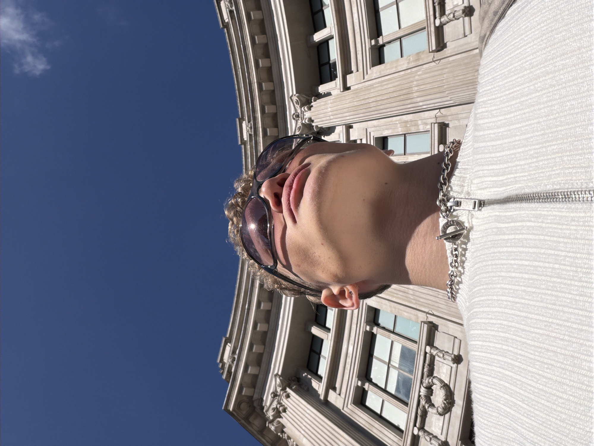 A low-angle photograph taken outdoors in the city of London, United Kingdom, features a person in sunglasses, wearing a silver chain necklace and a white textured jacket. The subject's face, especially the nose and lips, is prominently displayed. Behind the person, a portion of a light-colored, ornate building is visible, with windows and architectural details, set against a clear blue sky with a single cloud.