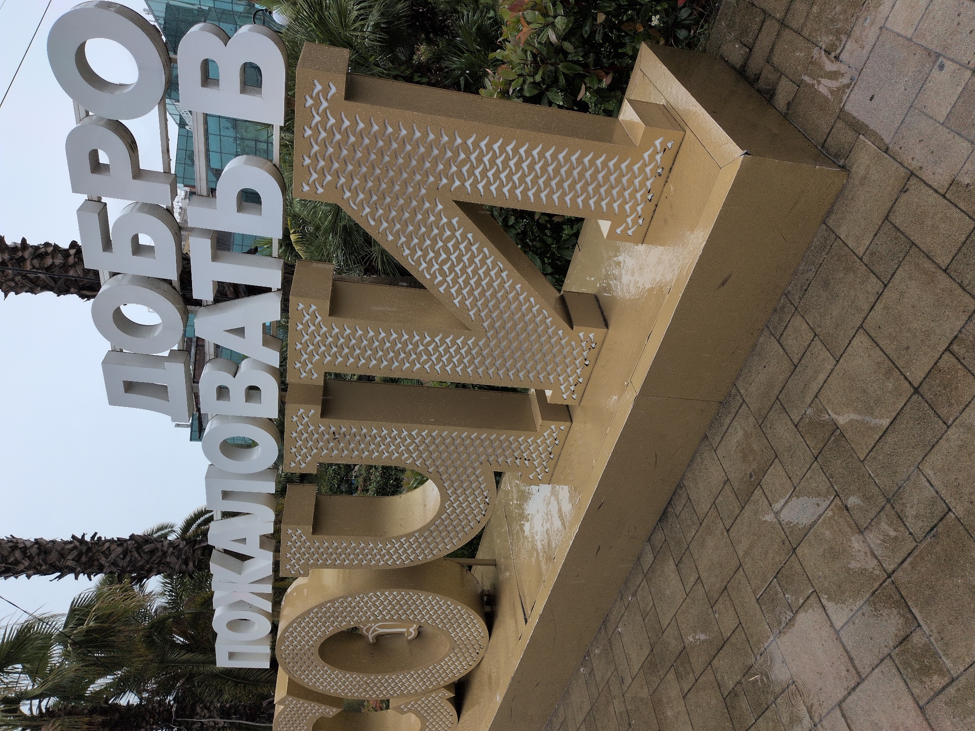 The image depicts a large, golden, three-dimensional sign in an outdoor setting. The sign prominently features text in Russian, with "ДОБРО ПОЖАЛОВАТЬ В СОЧИ" visible. The letters are large and stylized. The foreground shows a wet, paved walkway, suggesting recent rain. In the background, there are palm trees and a modern building with a blue facade. The sign appears to be a welcome sign for the city of Sochi, Russia, a popular resort city on the Black Sea coast. The overall impression is one of an entrance or prominent landmark in a tourist area. There are no people visible in the image.