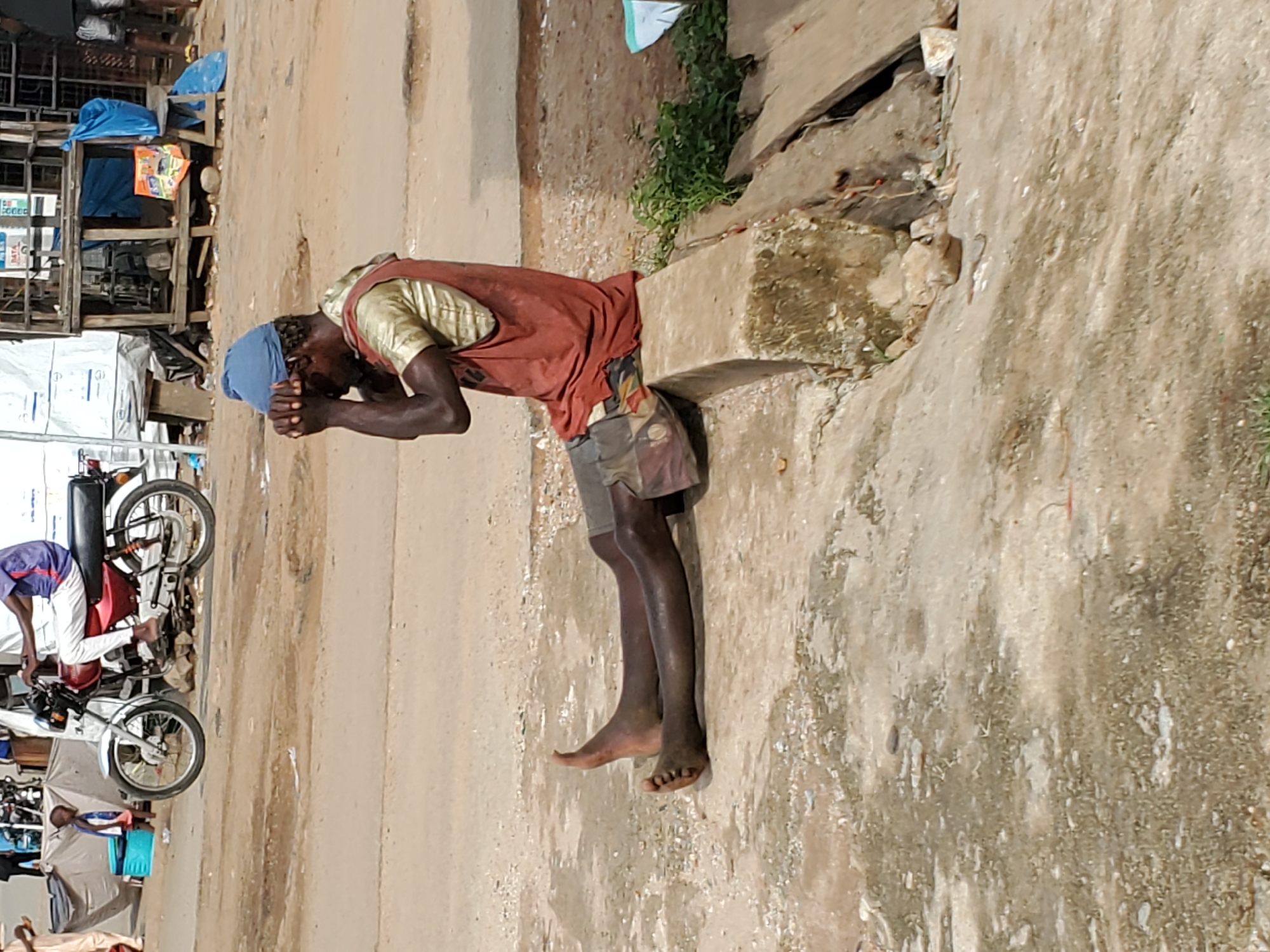 An outdoor scene in Mararaba, Nigeria, captures a moment of solemnity or distress. In the foreground, a dark-skinned man is positioned, bent over a concrete structure with his hands clasped near his face, suggestive of prayer, meditation, or perhaps exhaustion. He wears a blue head covering, a light-colored, possibly gold or light green, short-sleeved shirt, and a reddish-brown sleeveless top or apron over it. His lower body is clad in patterned shorts, and he is barefoot, with his feet showing some dust.
The man is leaning against a rough, light-colored concrete block, part of a larger structure. Behind him, the ground is unpaved, dusty, and beige, with some sparse green vegetation growing from cracks. The background reveals a bustling street or market area. To the left, several motorcycles are visible, one with a person seated on it, and others parked. Simple wooden stalls or makeshift structures, some covered with blue tarpaulins or sheets, line the side of the road, suggesting vendor activity. The overall impression is one of an everyday scene in a busy, perhaps economically modest, urban or semi-urban environment.
The lighting is bright, indicating daytime with clear skies, casting distinct shadows. No legible text is visible on any signs or objects within the image. The scene conveys a sense of candid street photography, focusing on a personal moment amidst the daily activity.