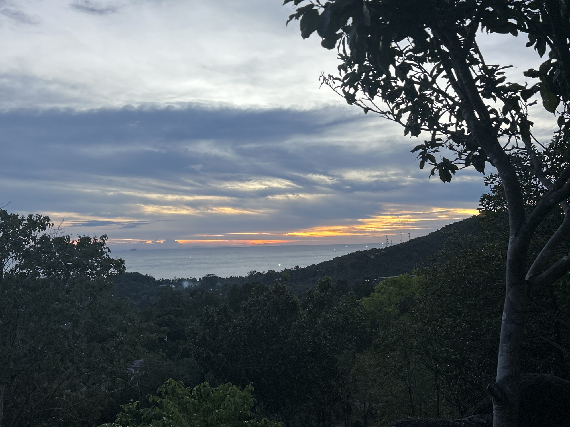 The image depicts a sunset over the ocean, viewed from an elevated position in Baan Hin Kong, Thailand. The foreground is dominated by dense green vegetation, including trees with dark green leaves. A tree with a light gray trunk and prominent branches frames the right side of the image. The middle ground shows a coastline and rolling hills covered in trees. The ocean horizon is illuminated by a strip of orange and yellow light from the setting sun. The sky is filled with layers of clouds, ranging from light gray to darker blue-gray tones. In the distance, several radio towers or antenna-like structures are visible on the horizon. No people are visible in the image.