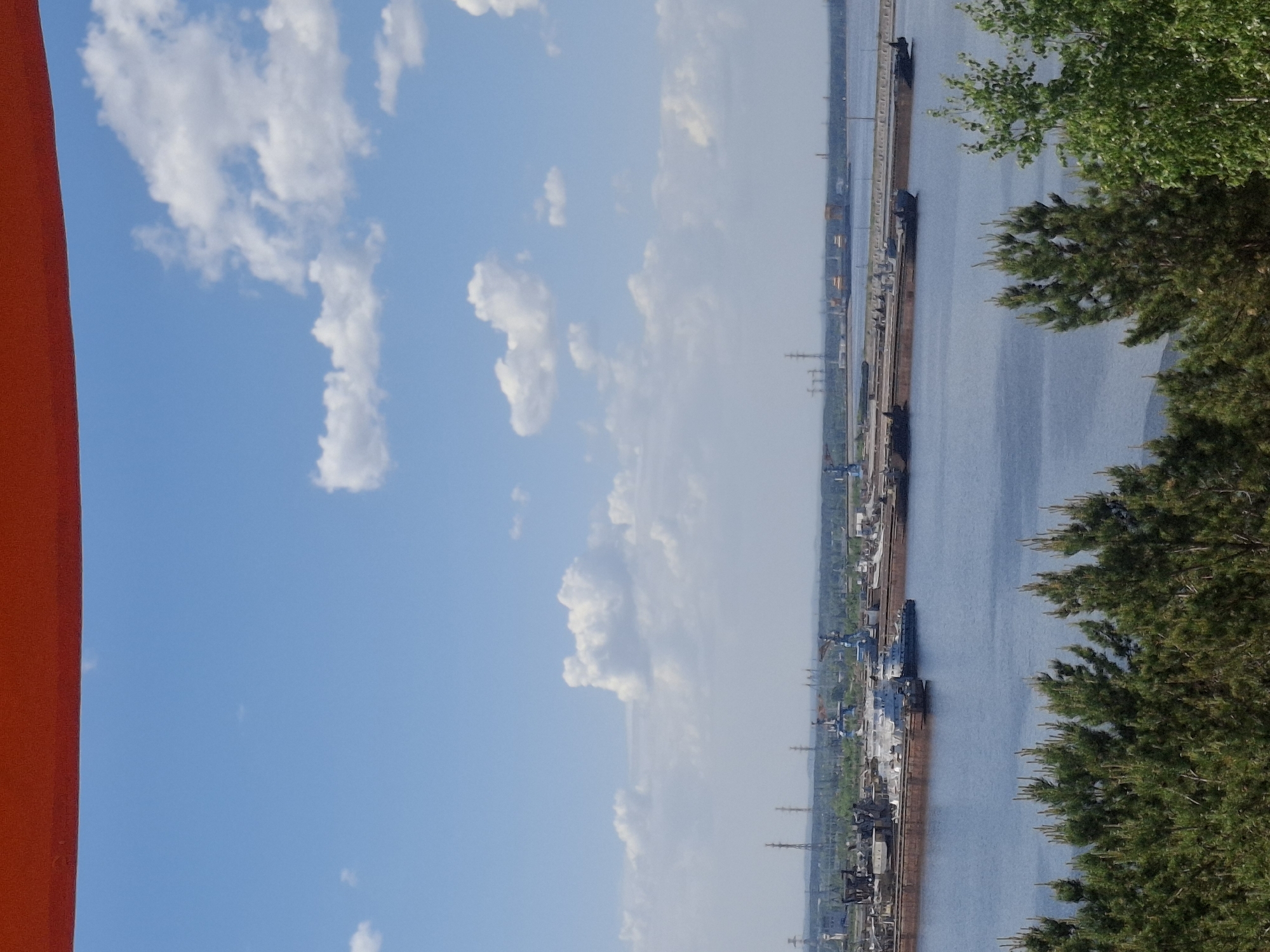 View from an elevated position in Chaikovsky, Russia, oriented vertically. A section of a red structure is on the left. A blue sky with white cumulus clouds spans the upper portion. Below, the Kama River stretches into the distance. The river is lined with a dock, where cargo ships are moored. An industrial zone runs parallel to the dock, with a series of pylons and industrial buildings. Foliage from nearby trees is visible on the right side.