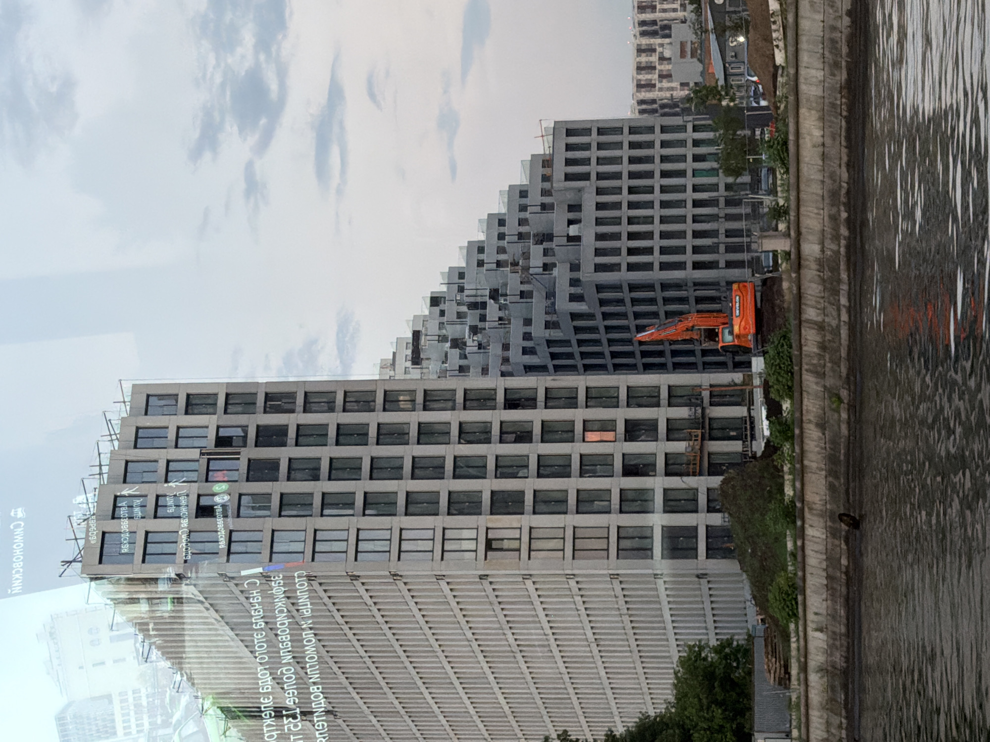 The image captures a view of a riverfront construction site in Moscow, Russia. In the foreground, the murky water of a river flows, with a dark object, possibly debris or a small animal, partially submerged. A concrete embankment lines the river, behind which is a construction site. Piles of dirt and vegetation are visible, along with stacks of wooden planks and construction fencing.
Dominating the background are two large, modern buildings. The building on the left is a tall, rectangular structure with a light-colored facade featuring vertical ribbed detailing and numerous windows. Overlaid on this building, and visible as a reflection or overlay, are reversed Russian characters, possibly from an advertisement or signage.
To the right of this building is a complex, stepped structure with a grey facade and many windows, appearing to be under construction. An orange excavator with the "DEVELON" logo is positioned on the construction site in front of this stepped building, its boom arm extended. Further to the right, another building with a patterned facade is visible in the distance. The sky is overcast with scattered clouds.