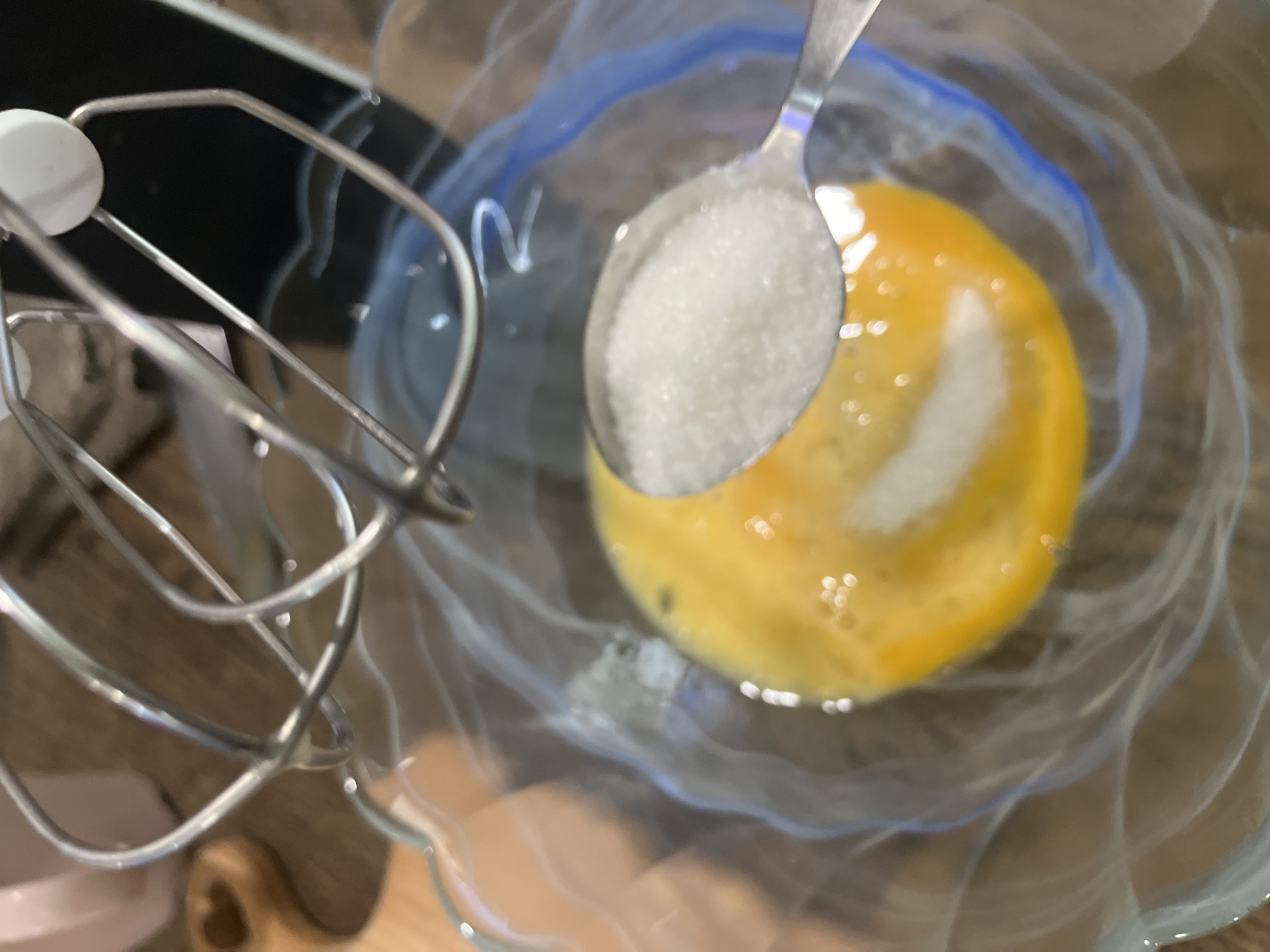 A close-up, high-angle shot depicts a culinary scene in progress. A clear glass bowl, rimmed with a blue design, contains an egg yolk and a spoonful of granulated white sugar. A metal spoon filled with sugar is poised above the yolk, presumably about to deposit its contents. To the left of the bowl, the metal beaters of an electric mixer are visible. The background is a blurred wooden surface. The image provides no information to determine the location in Krasnodar, Russia or to suggest that it is there.