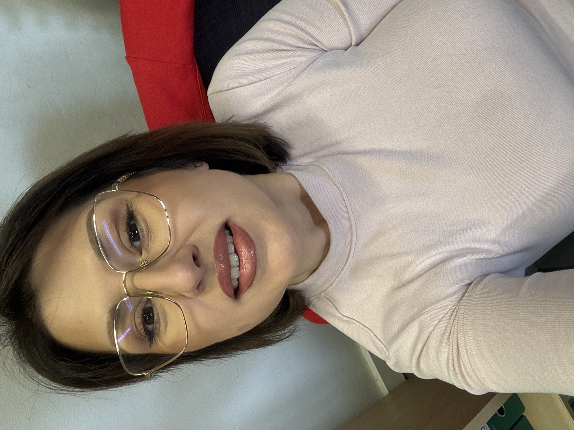 A medium shot of a woman with light skin, short dark brown hair, and wearing gold-rimmed glasses. She is looking directly at the camera with slightly parted, glossy lips. She is wearing a light pink long-sleeved shirt and sitting in a chair with red and black fabric. The background appears to be a light-colored wall and a wooden desk or shelf. The location is undetermined.