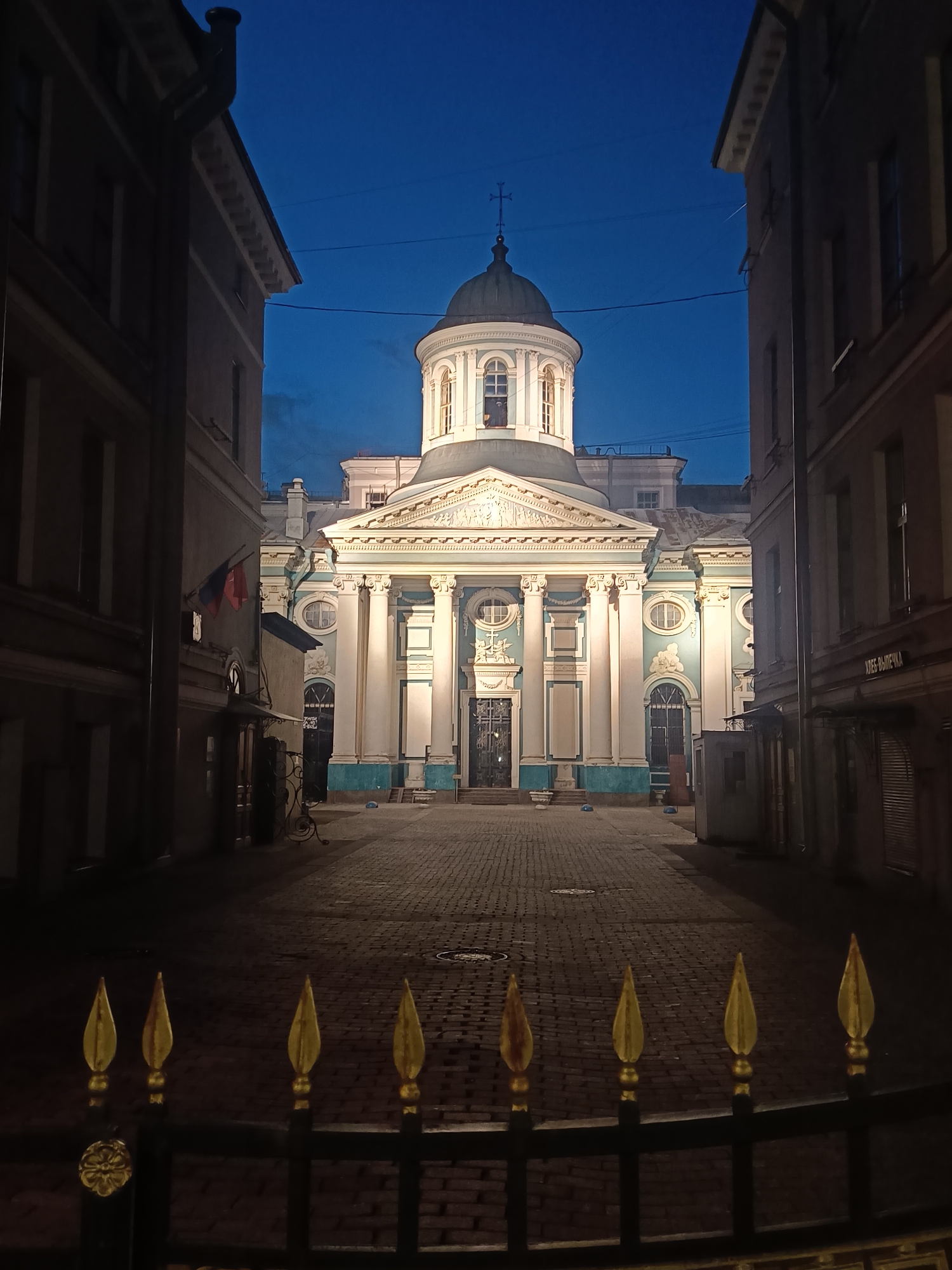 A nighttime scene depicts a clear view of St. Panteleimon Church in Saint Petersburg, Russia. The baroque architecture of the church is illuminated, emphasizing its columns, dome, and ornate facade. The church is framed by buildings on either side of a narrow street, adding to the depth of the composition. In the foreground, a black metal fence with gold spear-shaped finials adds a decorative element. The sky is a gradient of deep blues, suggesting the scene is captured during twilight or early evening. The cobblestone street leads directly to the church entrance. No people are visible in the shot.