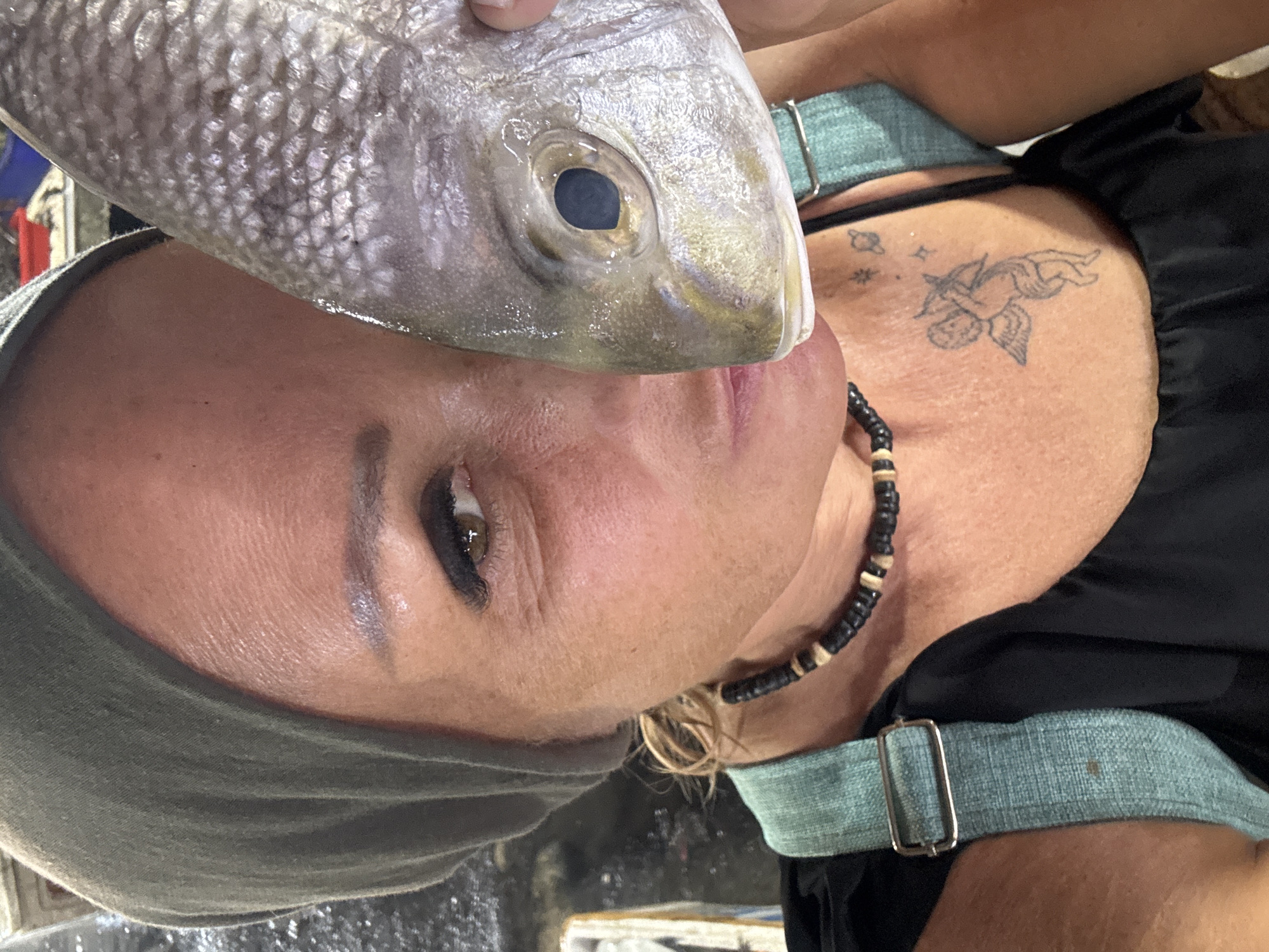 A woman wearing a green head covering and a black and tan beaded necklace is holding a fish to her face. She has a tattoo of a cupid on her left upper chest. The background is not clear enough to establish the location.