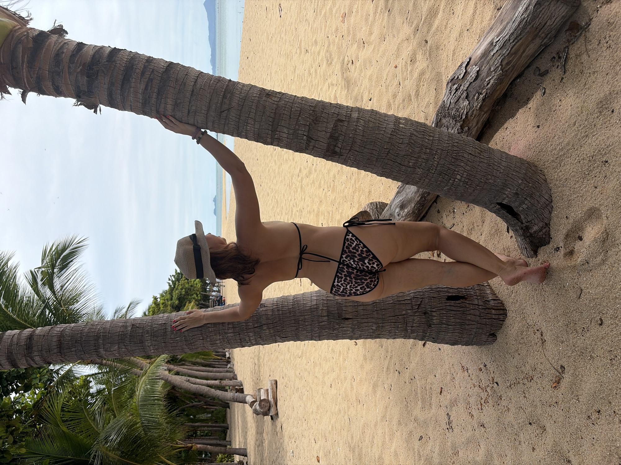 A woman in a leopard-print bikini and a straw hat is leaning against a palm tree on a sandy beach. She is holding onto the tree with her right hand, and her left hand is resting on the tree trunk. Her legs are crossed. There are other palm trees and foliage in the background, as well as the ocean and a clear sky. The sun appears to be out, suggesting a bright day. The scene is set at Baan Bo Phut, Thailand. The woman appears relaxed and enjoying her time outdoors. There is no visible text in the image.