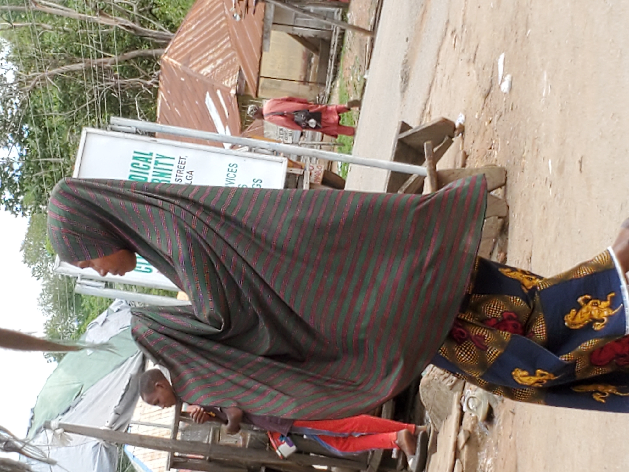 A medium shot shows a woman in Mararaba, Nigeria. She is wearing a dark green, horizontally-striped hijab with red stripes. Her dress is black with gold lion figures and red, round shapes. The woman is looking down. In the background, a man walks away, wearing a red top and dark pants. Buildings with rusted metal roofs are also in the background. Part of a white sign is visible, partially obscured. A boy is seen seated at the bottom of the image.