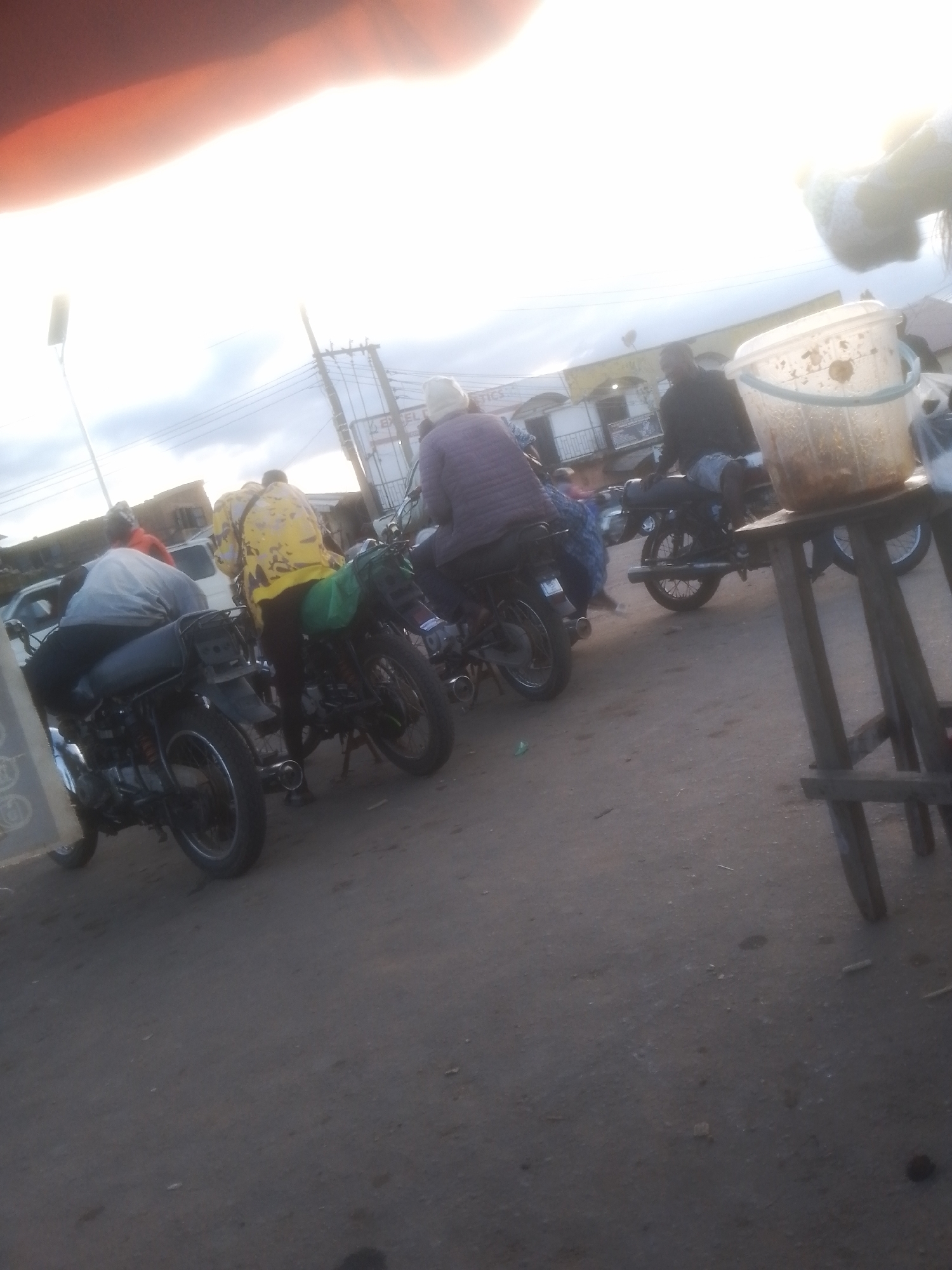 An outdoor scene depicts several people on motorcycles, suggesting a roadside gathering or transportation hub. In the foreground, a person wearing a grey shirt and black pants is seated on a motorcycle. To their right, another person in a bright yellow patterned shirt and black pants stands next to a motorcycle. Further in the background, a person in a purple shirt and white headwear is on a motorcycle, and another individual in a dark shirt and shorts is also on a motorcycle. A clear plastic bucket filled with food or liquid sits on a wooden stand to the right of the frame. The ground is paved, and overhead power lines are visible against a bright, cloudy sky. The overall impression is a busy, informal setting in an urban or semi-urban environment.
