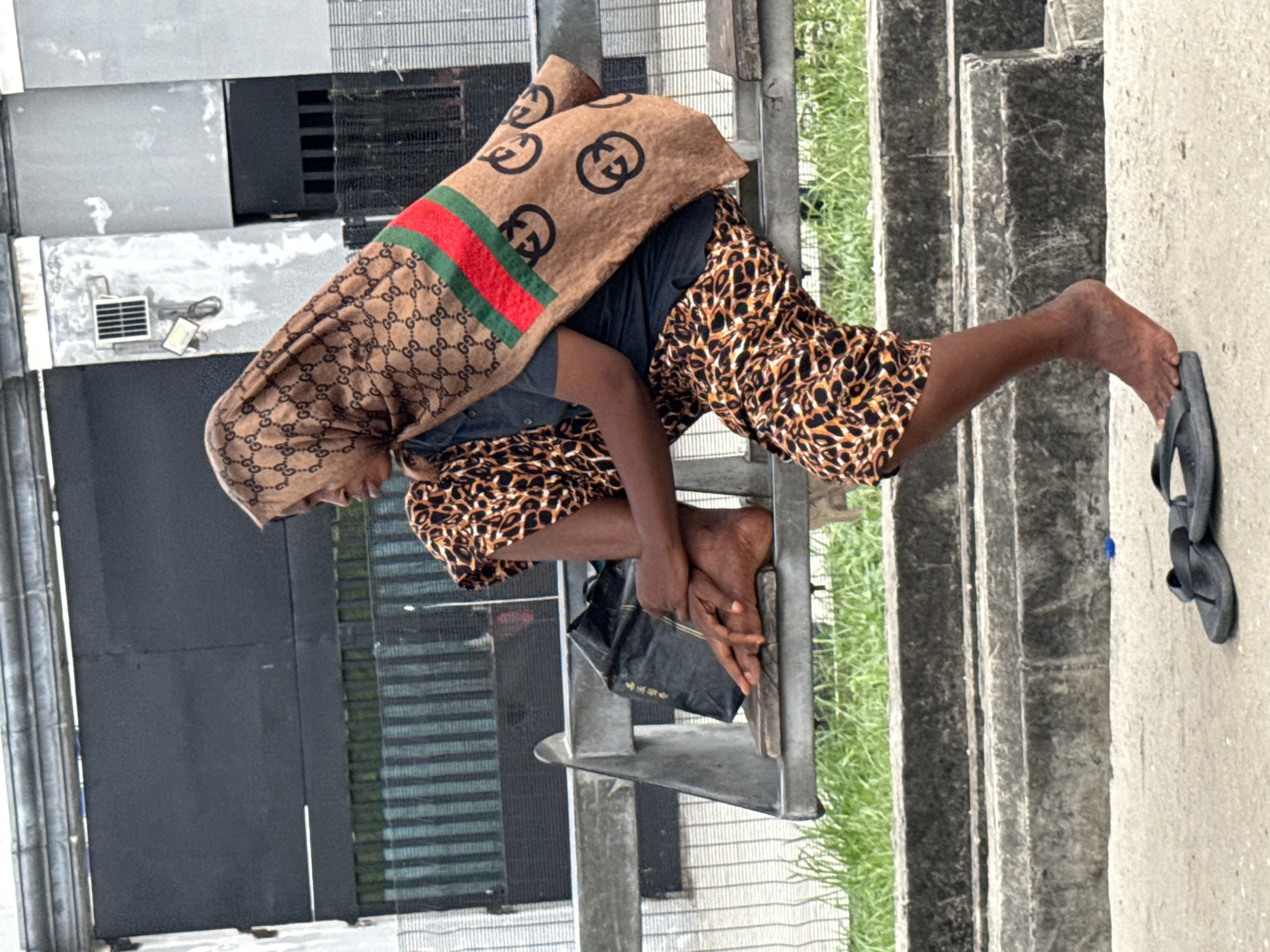 An individual in a brown, Gucci-patterned head covering and animal-print attire sits on a metal structure near concrete steps. Sandals are lying on the concrete next to the steps. The setting is Igboefon, Nigeria. A building with gray walls and a barred window is visible in the background. Greenery is visible to the side of the steps. The individual has crossed their legs and is holding a black object in their hands.