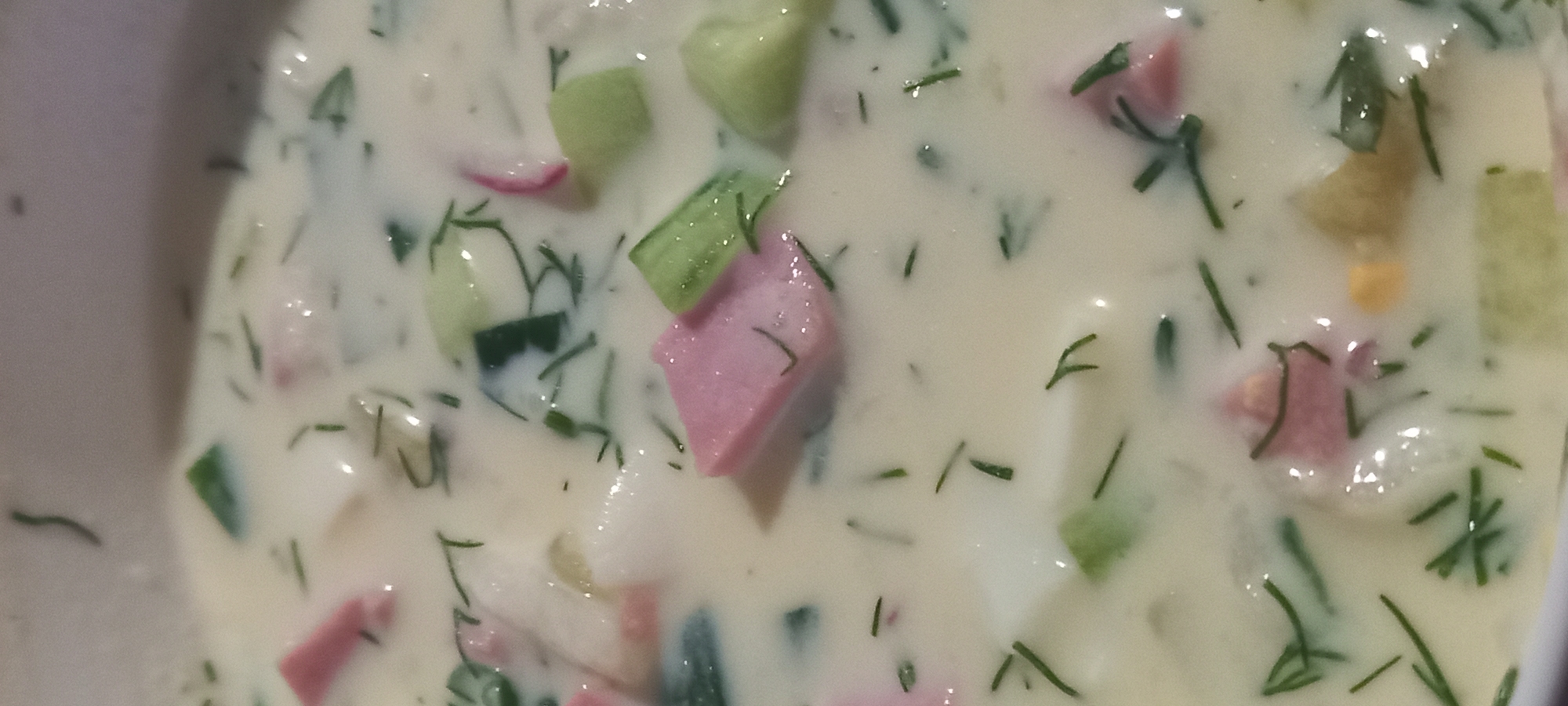 The image shows a close-up of a bowl of Okroshka, a cold soup. The soup is creamy white, with visible pieces of chopped vegetables such as cucumbers, radishes, and dill. There are also pieces of pink-colored ham or sausage. The lighting is dim, creating a soft focus effect. The image is a still life and does not depict any people or a specific location in Kryvyi Rih, Ukraine.