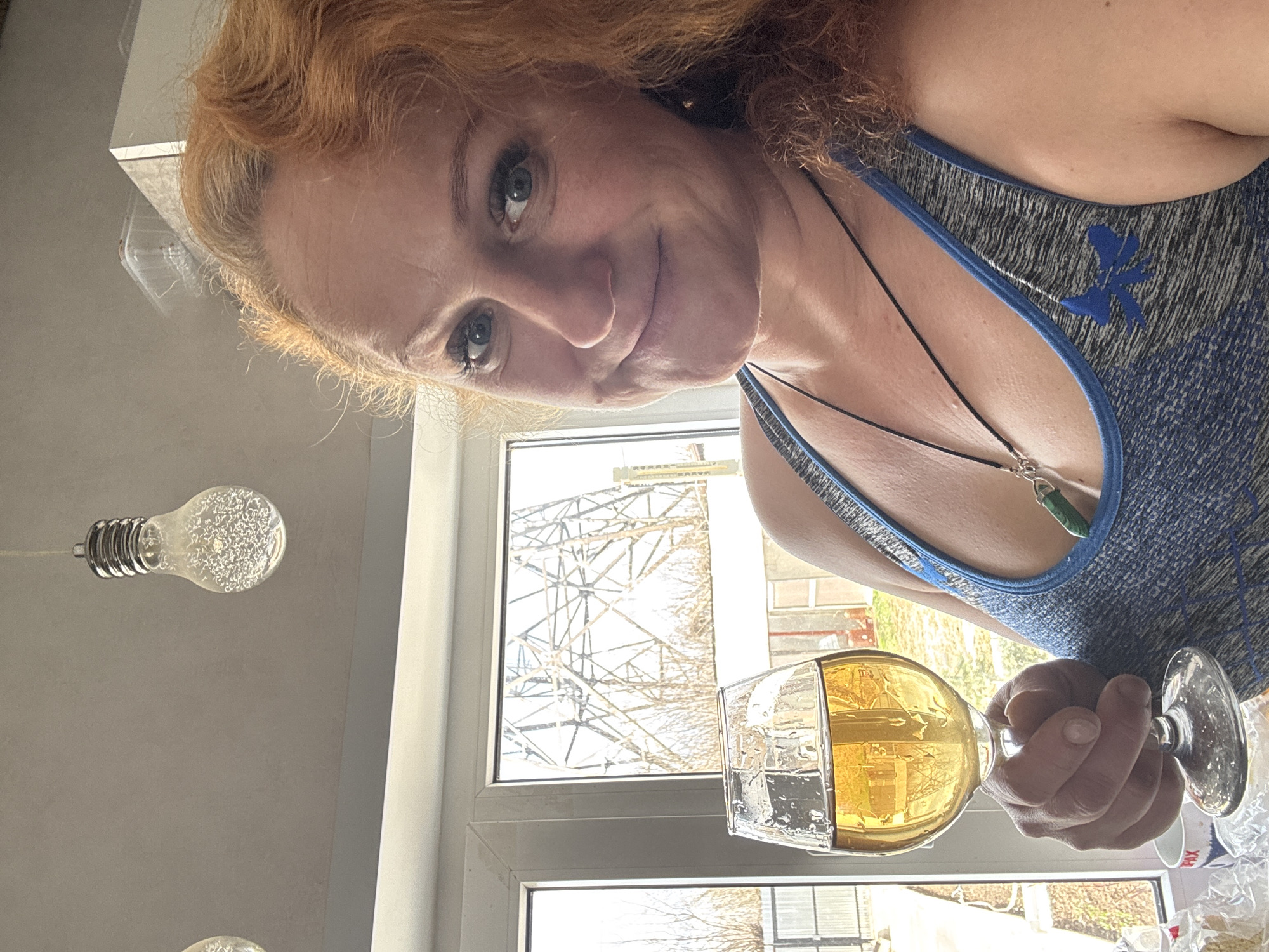 The media file displays a woman posing for a selfie indoors, likely in a kitchen or living room, with a window behind her. She has red or auburn, wavy hair and light-colored eyes, looking directly at the camera with a gentle smile. She is wearing a grey and blue athletic top or sports bra with a blue, stylized bird or leaf-like logo on the strap. A thin black cord necklace with a green, elongated pendant hangs around her neck.
In her right hand, she holds a stemmed glass containing a yellow/amber liquid, possibly wine, beer, or juice.
Behind her, a large window reveals an outdoor scene, suggesting daytime with bright natural light. The view through the window includes bare trees, indicative of winter or early spring in a temperate climate like Chaikovsky, Russia. A large metallic structure, possibly a power line tower or industrial frame, is visible through the upper part of the window.
To the left of the woman, two modern light fixtures featuring exposed, decorative filament-style bulbs are visible hanging from the ceiling. On a white object partially visible through the glass she is holding, the text "PAX" is discernible. The overall mood appears casual and relaxed.