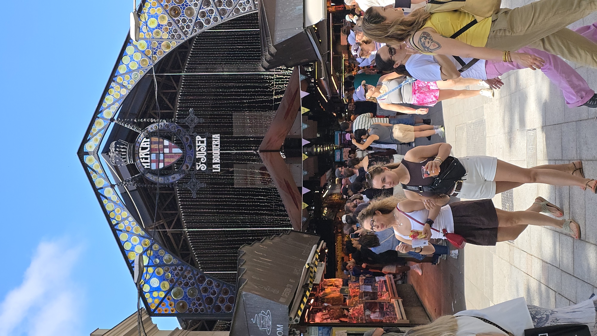The image captures a bustling scene at La Boqueria Market, a famous food market located on Las Ramblas in Barcelona, Spain. In the foreground, several women are walking on a paved pedestrian walkway. One woman on the left wears a white tank top, brown shorts, and sandals, carrying a red purse. Next to her, another woman in a dark tank top, white shorts, and sandals is holding a drink and a black crossbody bag. Further to the right, a woman in a yellow tank top and tan pants walks past with a backpack.
The background is dominated by the distinctive entrance archway of La Boqueria market, adorned with colorful mosaic tiles and a crest featuring the words "MERCAT S JOSEP LA BOQUERIA". Hanging strings of lights descend from the arch. In the mid-ground, a dense crowd of people browses the market stalls, which are visible on the left side of the image, displaying various food items. The overall atmosphere is lively and vibrant, typical of a popular tourist destination on a sunny day, indicated by the bright light and clear blue sky.