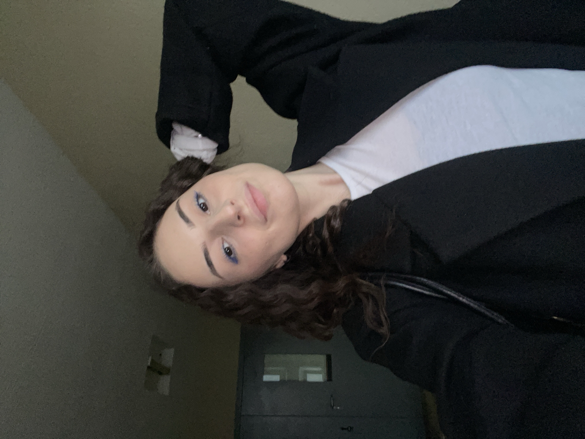 A young woman with dark curly hair and blue eyeliner is shown in a selfie. She is wearing a white top under a black coat. A closed door and a light switch are visible in the background, suggesting an indoor environment. No specific details indicate this image was taken in Moscow, Russia.