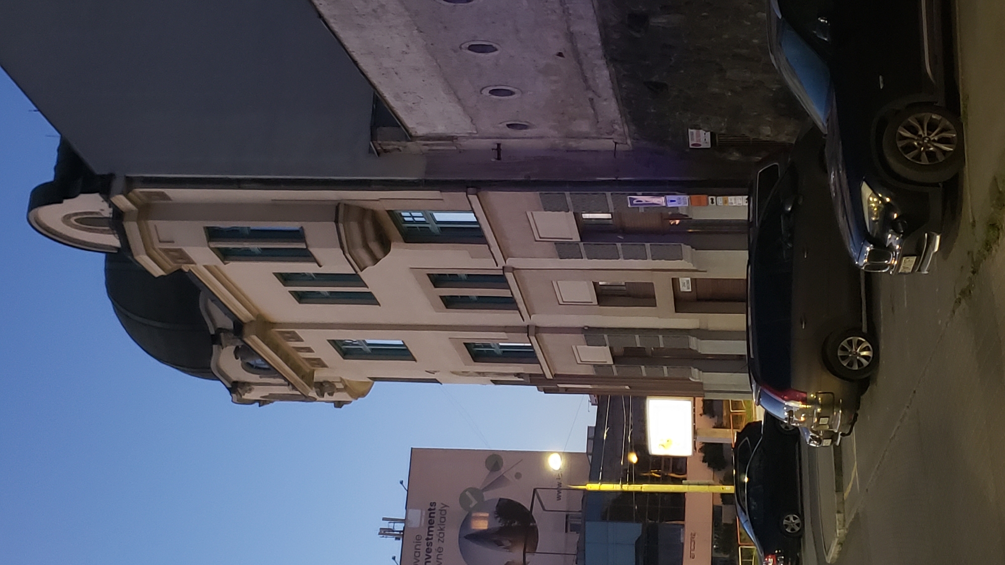 The image depicts an urban street scene in Prešov, Slovakia, likely during twilight or early evening. The focal point is an older, multi-story building with a distinctive domed roof and arched windows, showcasing architectural elements of a bygone era. Adjacent to it is a stark, gray, modern-looking building with a rough texture and a series of circular windows.
In the foreground, several vehicles are parked on asphalt, suggesting a public parking area. These include a dark sedan on the left, a darker-colored station wagon in the middle, and a black SUV on the right. The vehicles appear to be stationary.
In the background, a large billboard advertising "Investments" is visible, illuminated and featuring a circular graphic. A yellow lamppost stands to the left of this billboard. The sky is a deep blue, indicating the transition from day to night. The overall impression is one of a typical urban environment with a mix of historical and contemporary architecture and the presence of everyday street life.