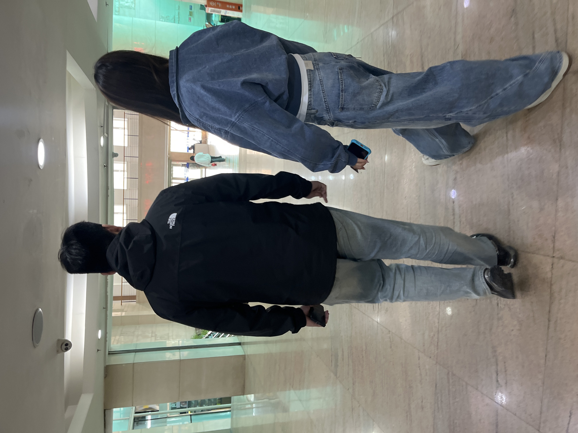 Two individuals are standing inside a building. The person in the foreground is wearing a black jacket, light blue jeans, and black shoes. The person in the background is wearing a blue denim jacket, light blue jeans, and white shoes. Both individuals are holding cell phones. The building has tiled floors and glass walls. This is in the city of Changchun City, China.