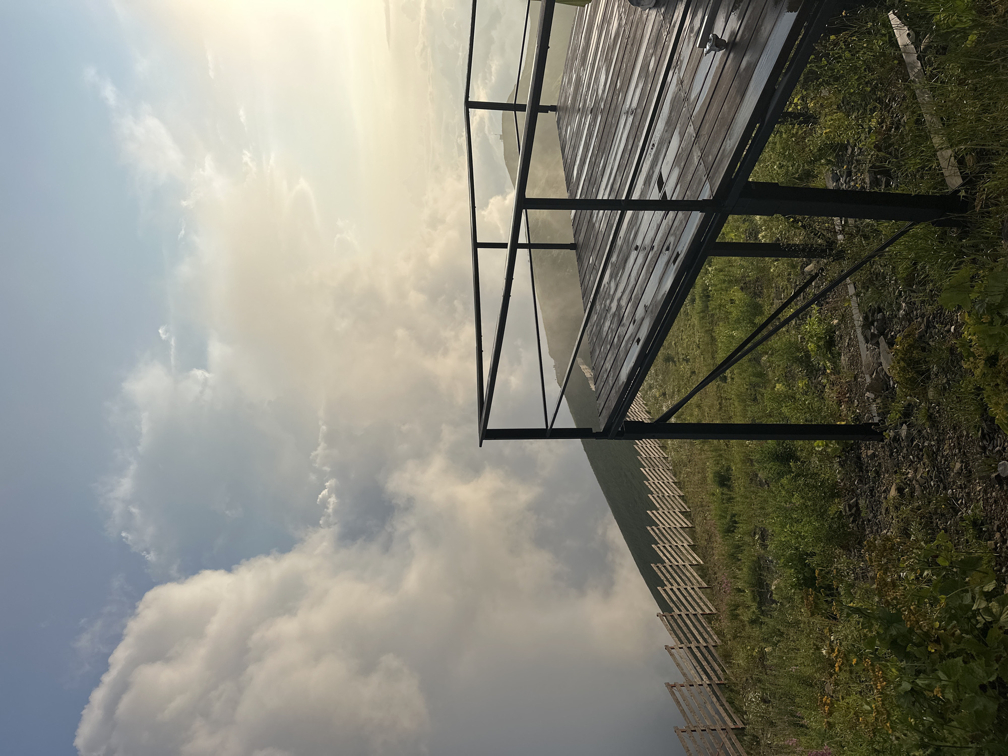The image shows an elevated wooden platform with a metal railing, positioned on a grassy mountainside. The platform appears wet, with puddles visible on the wooden planks. Beyond the platform, a wooden fence runs along the slope of the mountain. The sky is filled with large, dramatic clouds, some of which are illuminated by sunlight, suggesting a sunrise or sunset. In the distance, more mountains are visible, partially obscured by mist or clouds. This scene is characteristic of the mountainous terrain found in Esto-Sadok, Russia, a popular ski resort town located in the Krasnodar Krai region, near Sochi.