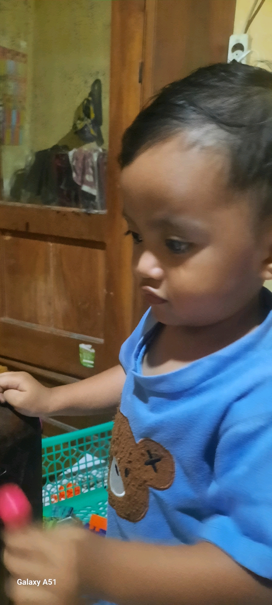 A medium shot captures a young child in a bright blue t-shirt with a bear design on it, positioned slightly to the right of the frame. The child's gaze is directed downwards and to the left. Their left hand rests on a dark surface, possibly a table, and their right arm is slightly blurred due to motion. The background consists of a wooden cabinet with a glass-fronted section that reflects light, revealing some items within. A green plastic basket is visible to the left of the child, containing colorful toy pieces. The environment appears to be indoors, with a warm, ambient light. The bottom left corner of the image displays the text "Galaxy A51". This image was taken in Jatitengah, Indonesia.