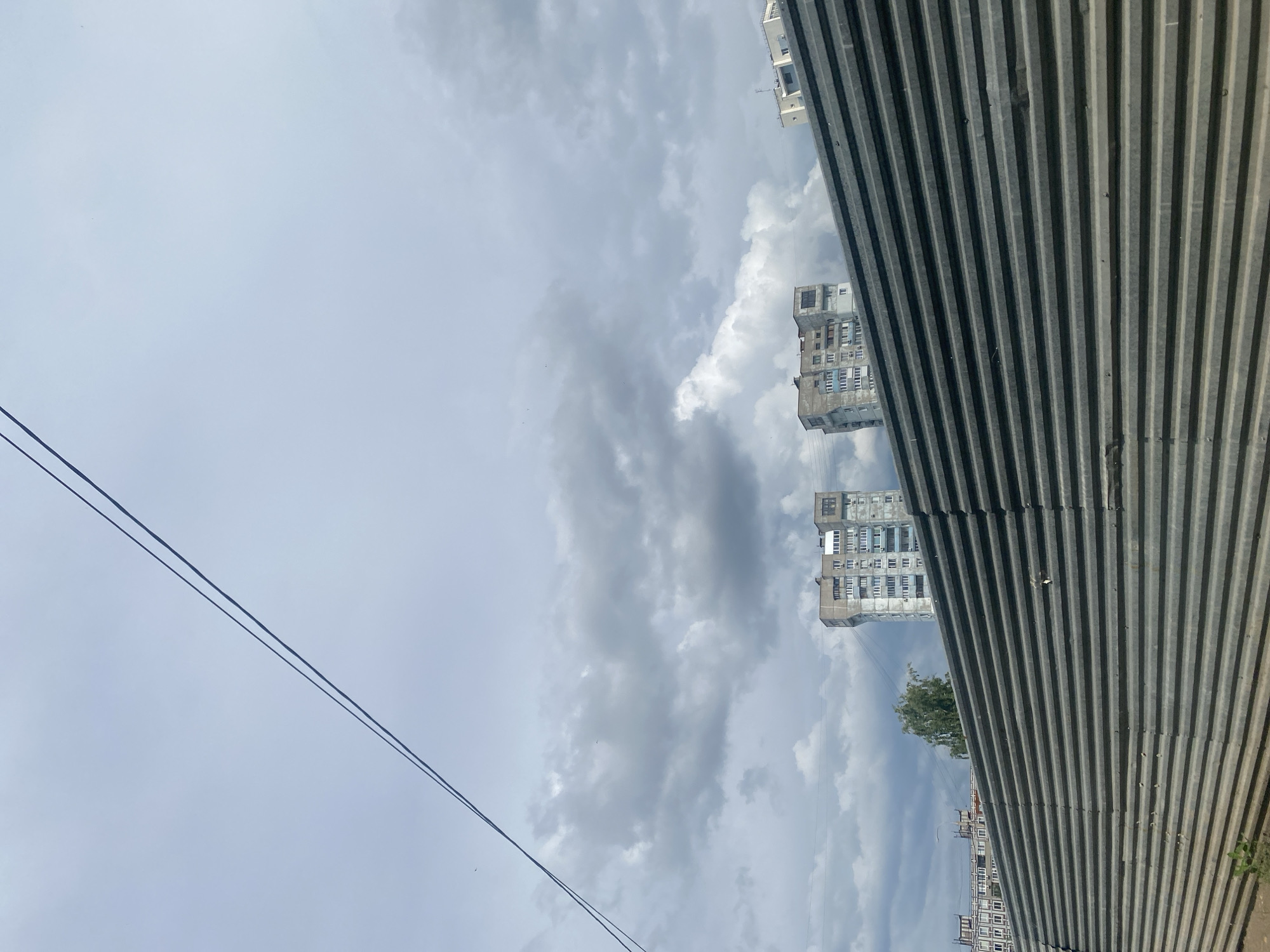 The image shows a view of a city skyline partially obscured by a corrugated metal fence in the foreground. Two multi-story apartment buildings, typical of Soviet-era architecture, are visible in the distance under a cloudy sky. The buildings have numerous windows and balconies. There are power lines running diagonally across the upper portion of the image. The fence is dark gray and appears to be made of corrugated metal sheets. The environment suggests an urban setting, possibly a construction site or an industrial area, given the fence. The location is identified as Samara, Russia. There are no people or animals visible in the frame.
