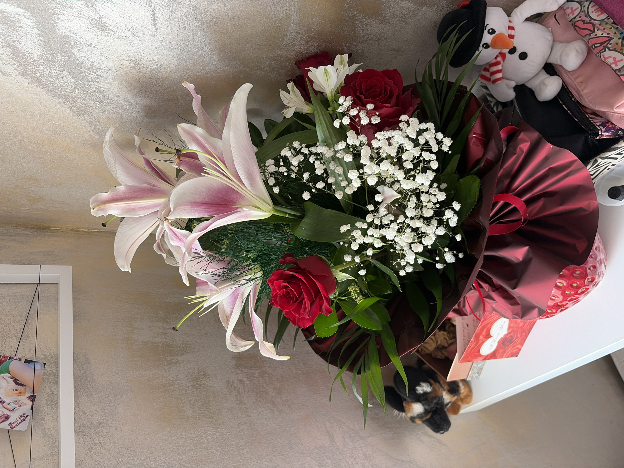 An arrangement of lilies and red roses is displayed on a white shelf in an indoor setting. The bouquet is wrapped in dark red paper and is adorned with a red ribbon. Next to the bouquet, on the left side of the shelf, is a small stuffed dog. To the right of the flowers, a plush snowman wearing a black hat and a striped scarf sits on top of several bags. In the background, a framed picture hangs on a textured wall. The image does not provide specific details about the city of Burgas, Bulgaria, or any actions or interactions.