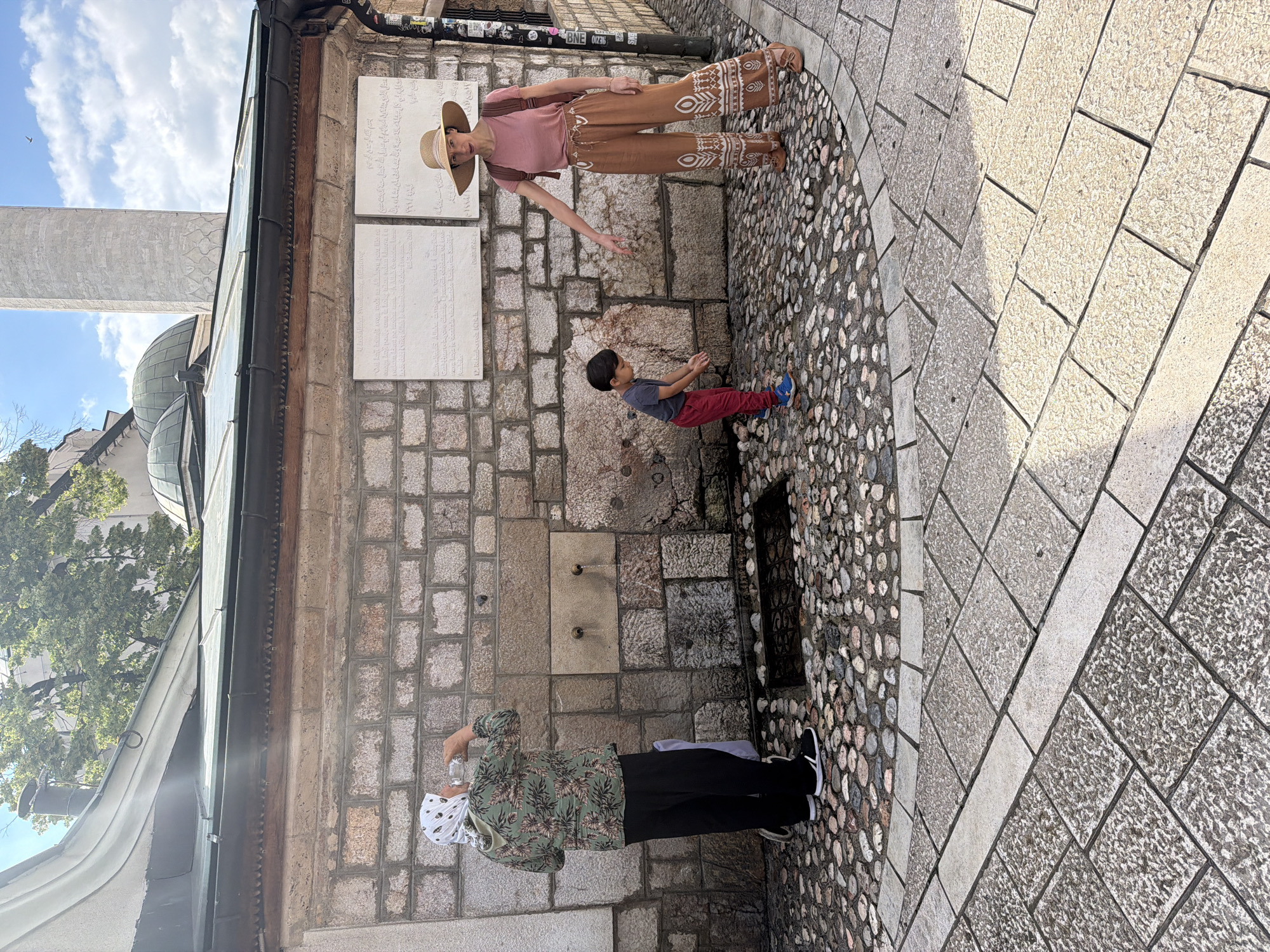 In Sarajevo, Bosnia and Herzegovina, outside a historic building constructed of rough-hewn stone blocks, three individuals are present near an outdoor water fountain. A person wearing a patterned headscarf, green floral-print jacket, and black pants stands on the left, drinking from a clear bottle. To the right, a young boy in a grey t-shirt and red pants is bending down, appearing to interact with the water. A woman in a pink t-shirt, a wide-brimmed hat, and patterned brown pants stands behind the boy, pointing towards the ground. The ground is paved with cobblestones. Behind the individuals, a stone wall features two white plaques with inscriptions, and a historical minaret rises in the background under a partly cloudy sky.