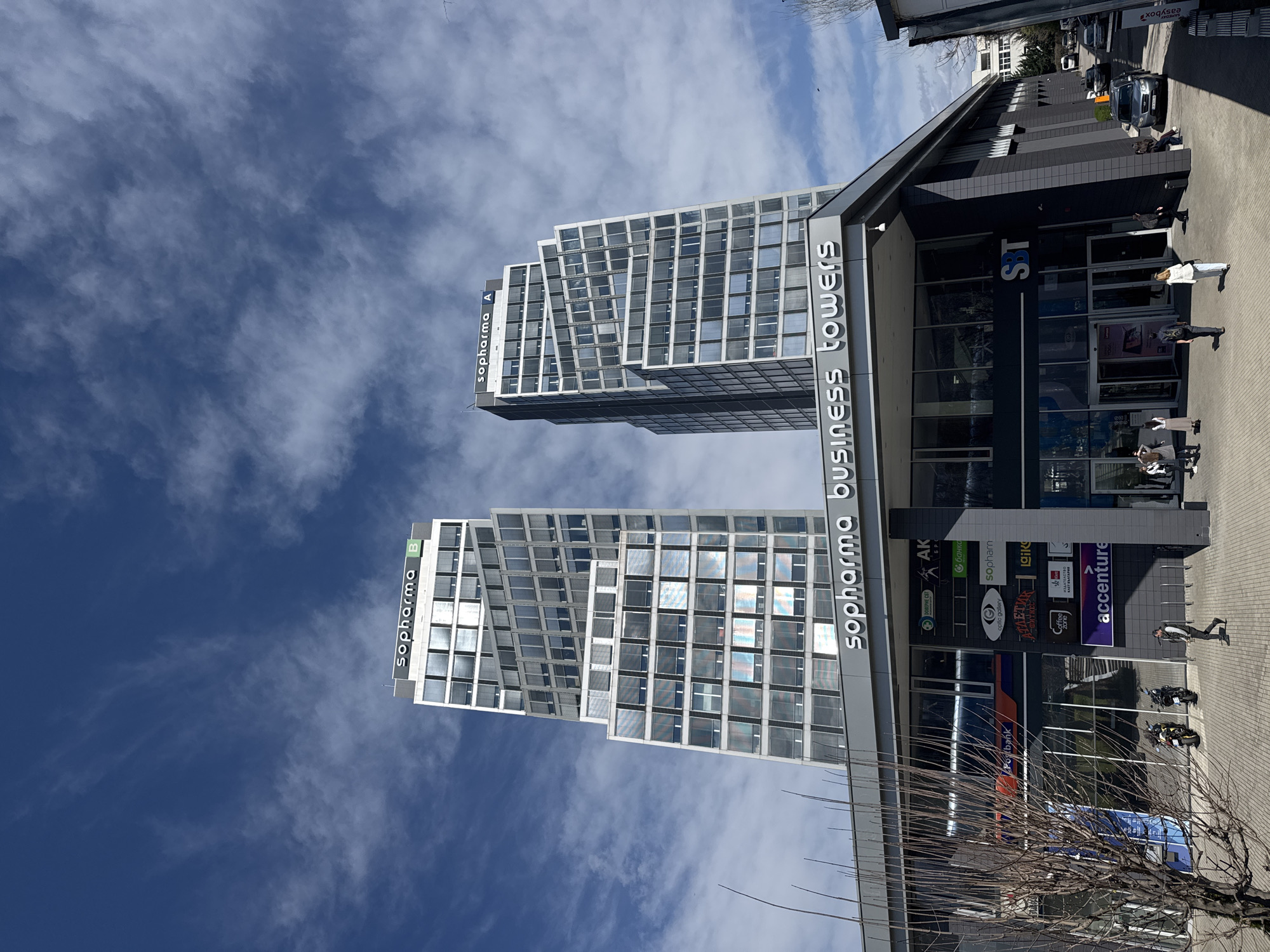 The image shows the Sopharma Business Towers in Sofia, Bulgaria. Two modern, high-rise buildings with glass facades are visible against a blue sky with scattered clouds. The base of the building complex features commercial spaces with storefronts including Accenture, with logos displayed. People are walking on the sidewalk in front of the building. Cars are parked on the side of the street. Bare tree branches are in the foreground. The sign “sopharma business towers” is prominently displayed on the building.