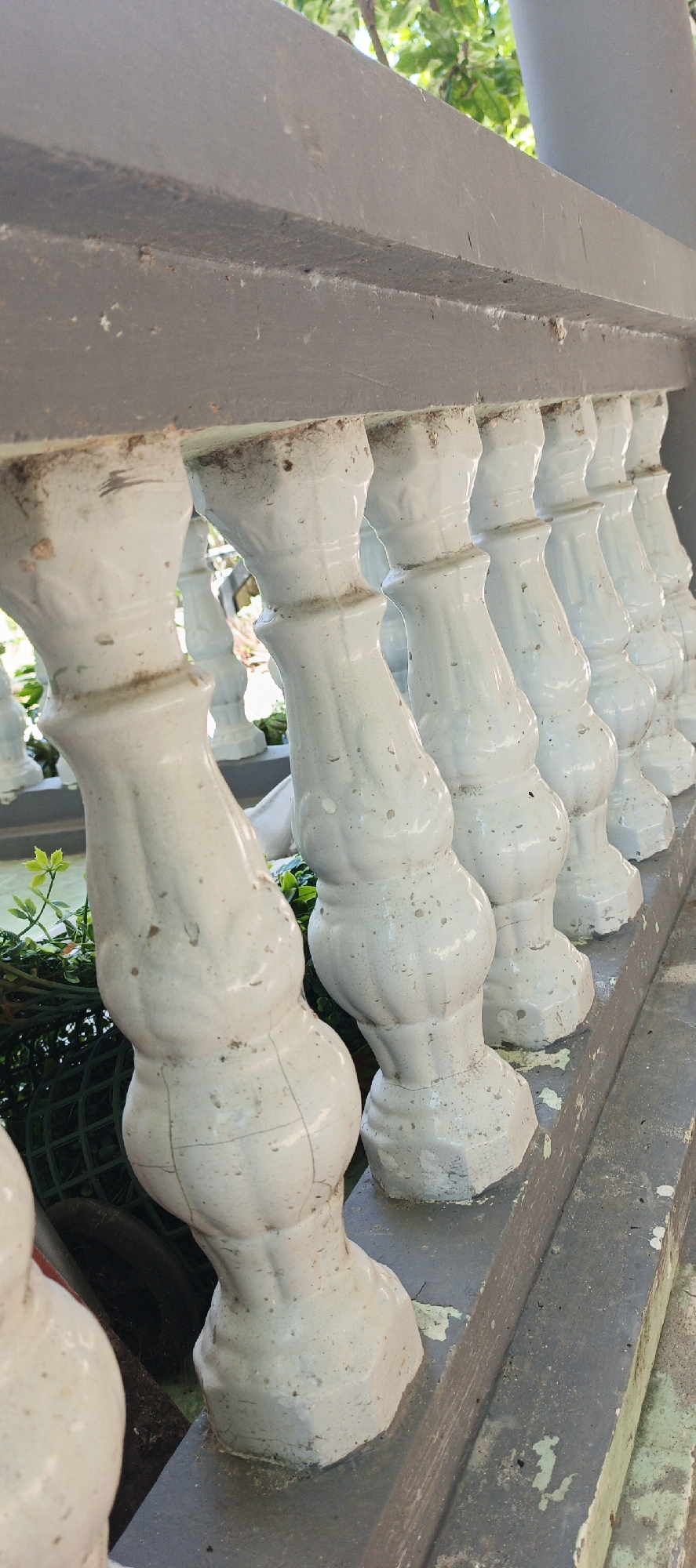 The image shows a close-up, eye-level shot of a weathered white concrete balustrade, likely part of a balcony or walkway, in Phan Thiet Ward, Vietnam. The balustrade features a series of decorative, turned posts with bulbous sections and carved details, arranged in a receding line. The concrete is chipped, stained, and shows signs of age, with dirt and debris accumulated in crevices. Behind the balustrade, lush green foliage and what appears to be a grey cylindrical pillar are visible, suggesting an outdoor setting. The top of the balustrade is a wider, grey concrete beam, also showing wear and tear. The overall impression is one of aged, outdoor infrastructure in a tropical environment.