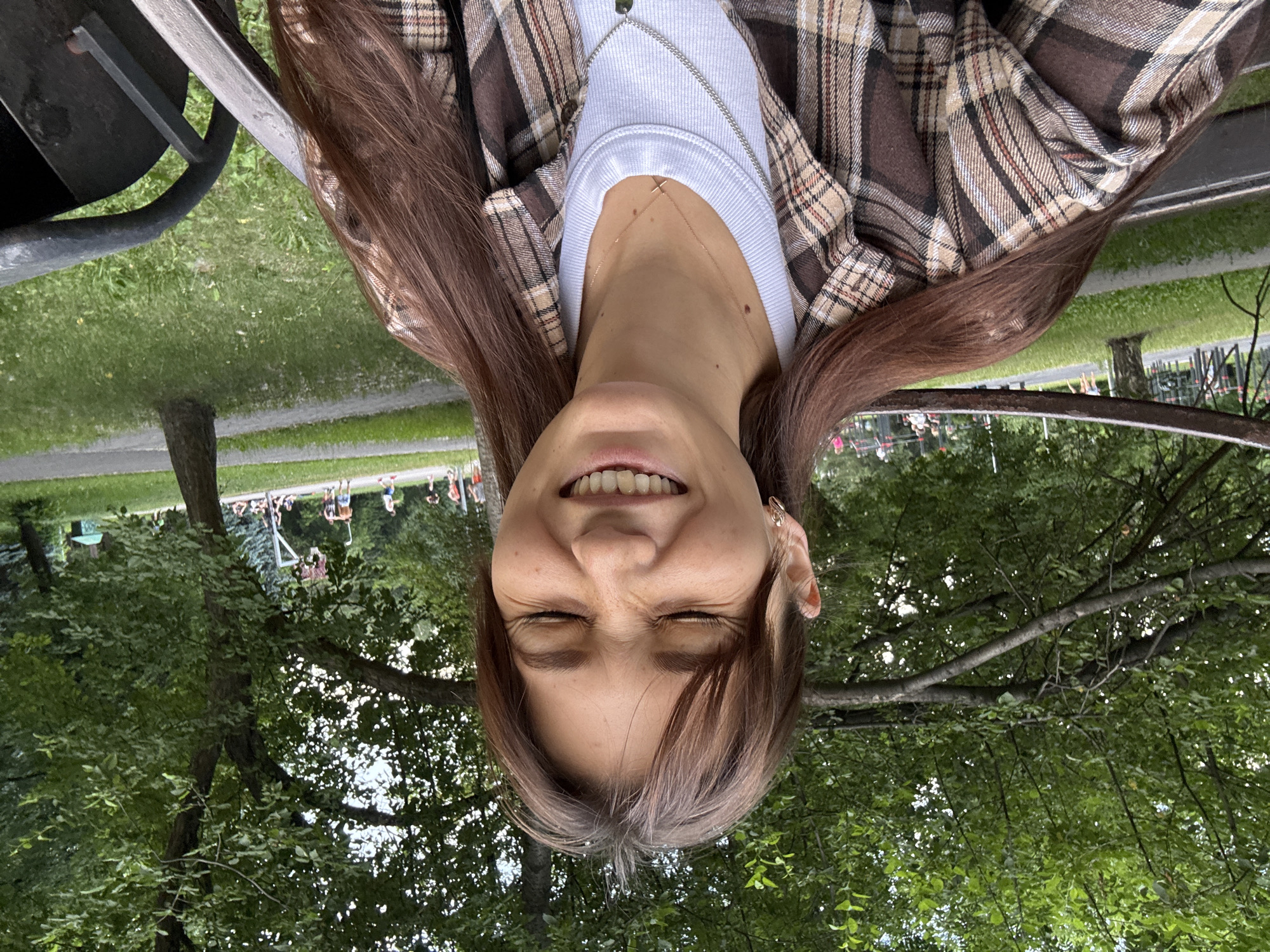 A young woman with long, dark brown hair and closed eyes is smiling in an outdoor setting. She is wearing a plaid shirt over a white t-shirt and two necklaces. She is sitting on a bench, with trees and greenery in the background. In the distance, people can be seen near a basketball court and playground equipment. The overall atmosphere suggests a casual day in a park. The location is a park in Novokuznetsk, Russia.