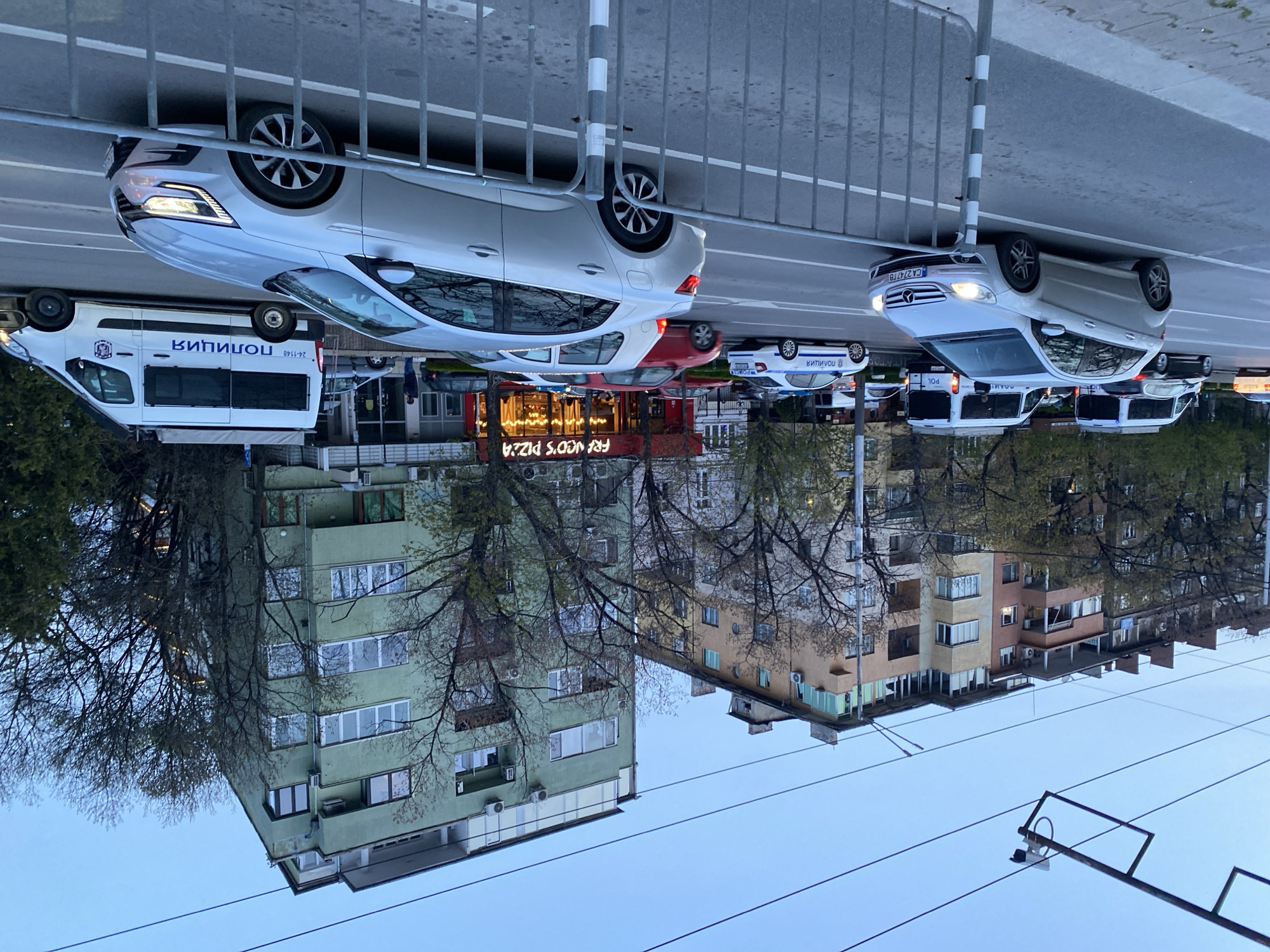 The image shows a street scene in Sofia, Bulgaria. In the foreground, two silver cars are parked near a metal railing. Behind them, a white van with "ПОЛИЦИЯ" (Police) written on its side is visible, along with a red building with "FRY N’ GO’S PIZZA" written on its front. Several other vehicles are parked further down the street. Buildings with residential apartments can be seen in the background with trees with few leaves. The sky is light blue. There are utility wires overhead.