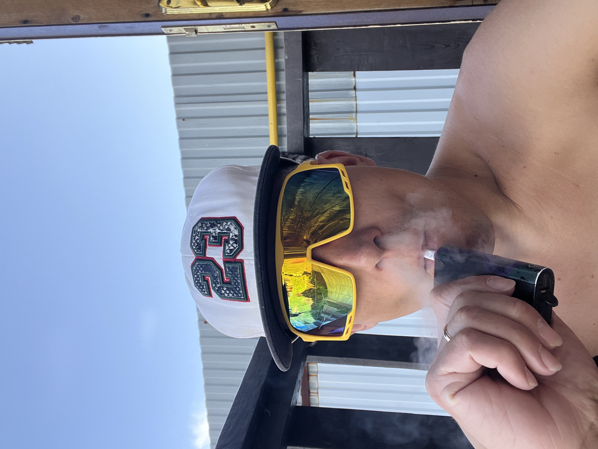 A person with a fair complexion, wearing a white baseball cap with the number "23" and yellow-framed sunglasses, is holding a vaping device and exhaling vapor. They are positioned in front of a corrugated metal structure with black wooden beams. The reflection suggests the sky and possibly trees are visible in the lenses of the sunglasses. Based on the location listed in the prompt, the picture was taken in or near Chaikovsky, Russia.