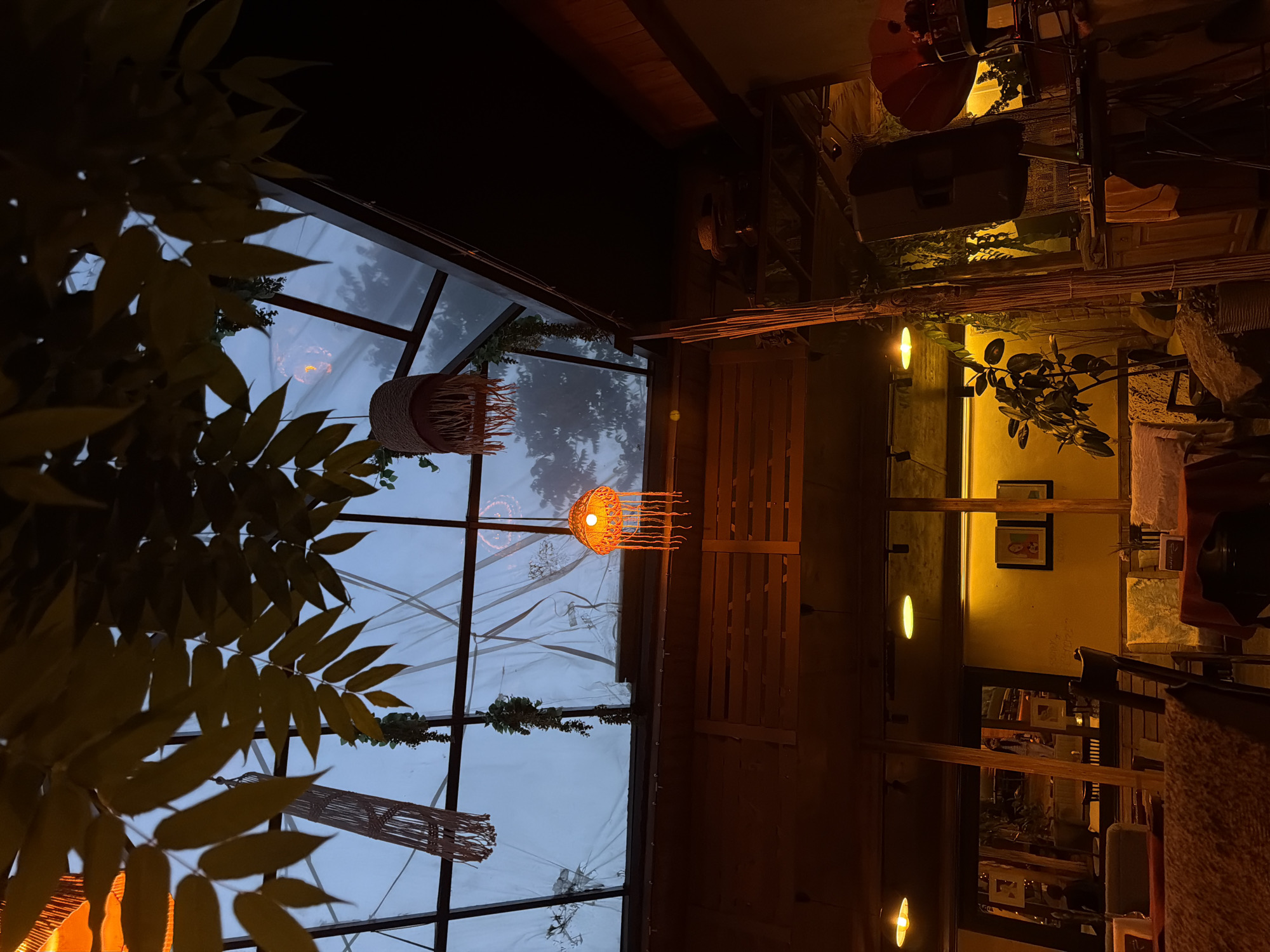 The image depicts the interior of a building in Yerevan, Armenia at night. A glass wall frames a view of a dark, overcast sky. Plants are visible near the glass wall. Inside, a wicker pendant light hangs from the ceiling. A wooden bench is positioned parallel to the glass wall. In the background, a yellow wall is illuminated by several small lights. Two framed pictures are displayed on the wall. Miscellaneous objects, including furniture and equipment, are visible in the background. The overall lighting is dim and warm.