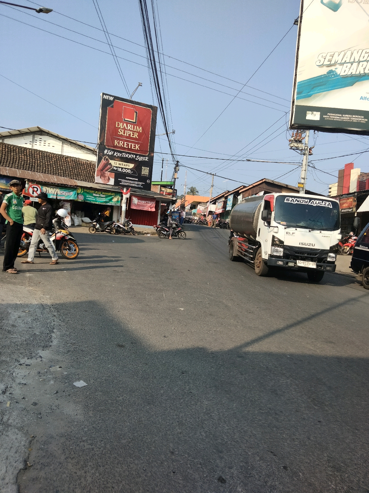 This image captures a street scene in Sawahan, Indonesia. In the foreground, a gray asphalt road shows signs of wear and tear. To the left, two men stand on the side of the road, one wearing a green t-shirt and black pants, the other in a black jacket and light-colored pants. A motorcycle is parked next to them.
In the middle ground, several other motorcycles are visible. A white Isuzu Elf truck with a tank body is driving on the road. A large billboard for "DJARUM SUPER KRETEK" stands prominently on the left side of the street. Behind the billboard, there are buildings with traditional tiled roofs. The background reveals more buildings, power lines crisscrossing the sky, and a clear blue sky. The overall atmosphere suggests a busy street in an Indonesian town.