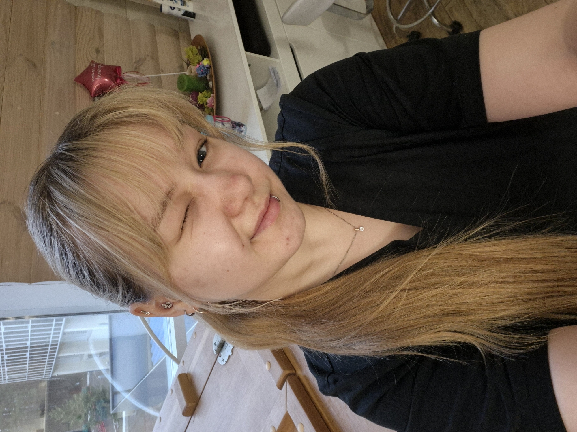 A young woman is captured in a selfie, winking playfully with her right eye and smiling slightly. She has long, straight blonde hair with darker roots, featuring bangs across her forehead. She wears a black t-shirt or top and a delicate silver necklace with a small, clear pendant. Multiple silver piercings adorn her left ear, including a small flower-shaped stud and several hoops. A silver hoop lip piercing is also visible on the right side of her mouth.
The setting is an indoor space, likely a home or room, with natural light streaming in from a window visible on the right side of the frame. The background includes various wooden surfaces, possibly part of a table, floor, or furniture, and hints of white shelving with some items on them. In the upper left background, a small, red, star-shaped foil balloon is visible. Next to it, a decorative arrangement in a wooden dish holds colorful artificial flowers in hues of green, blue, pink, and yellow, alongside a green cylindrical object.
The overall mood is casual and friendly. The image appears to be taken during the daytime, indicated by the natural light. Text is present on the red balloon, but it is blurry and unreadable. The interior scene is consistent with a residence, potentially in Chaikovsky, Russia.