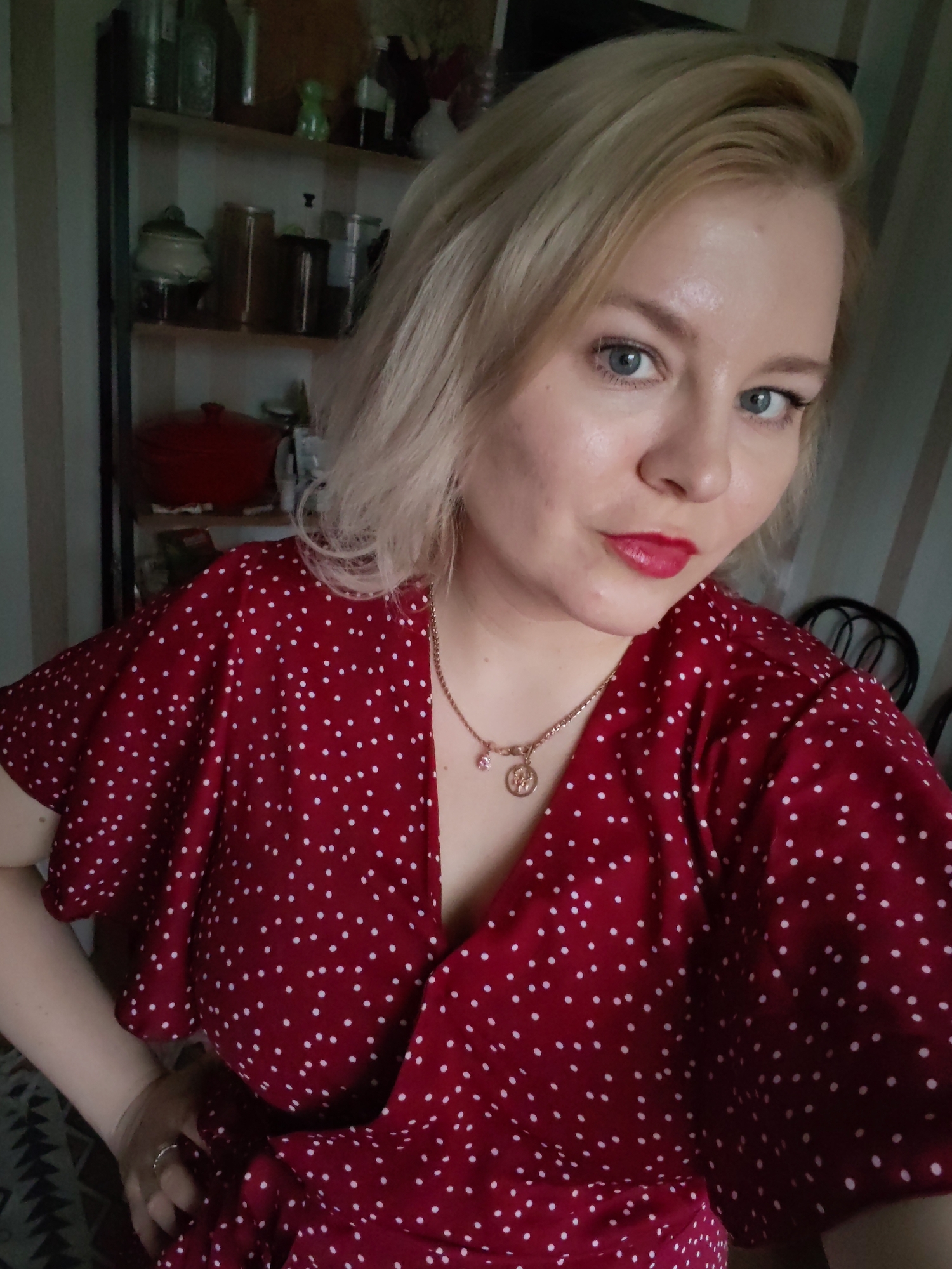 A medium close-up, slightly high-angle selfie portrait features a young adult woman with fair skin, light blonde hair, and blue eyes. She wears a vibrant red V-neck dress or top with white polka dots and short, fluttery sleeves. Her lips are accentuated with red lipstick, and she has a gold-colored necklace adorned with a pink charm and a circular medallion. A silver-toned ring is partially visible on her left hand.
The woman looks directly into the camera with a subtle, pleasant expression. The background indicates an indoor setting, likely a home in Izhevsk, Russia. To her left, a dark-framed shelving unit is visible, holding various household items including glass bottles, jars, a small light green decorative piece, and a prominent red lidded pot on the bottom shelf. The wall behind her features vertical light and dark stripes. A dark, ornate metal chair back is partially visible on the far right. The lighting suggests daytime. There is no visible text.
