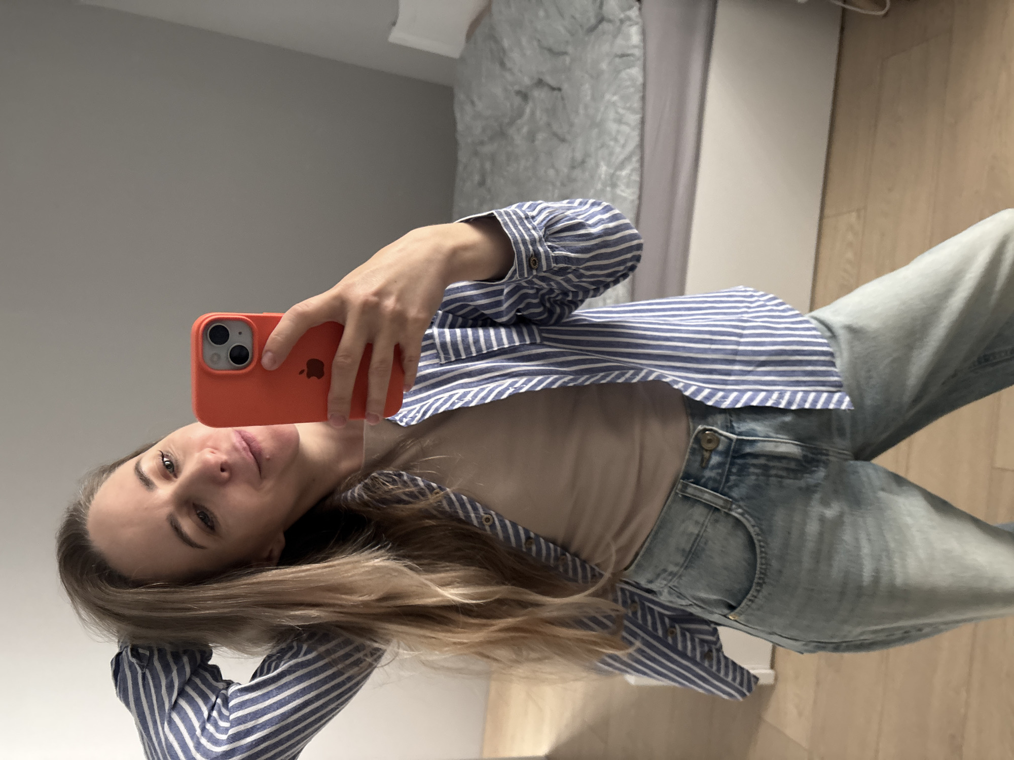 A woman is taking a selfie in what appears to be an indoor environment, possibly a bedroom. She is holding an orange iPhone in her right hand, capturing her image in a mirror. She is wearing a blue and white striped long-sleeved shirt, which is unbuttoned revealing a beige undershirt. She is also wearing light blue jeans. Her long blonde hair falls across her shoulder and towards the camera. In the background, a bed with grey bedding is visible, along with a light-colored wall and what appears to be a wooden floor. The photo does not contain enough information to determine if the location is Chaikovsky, Russia.