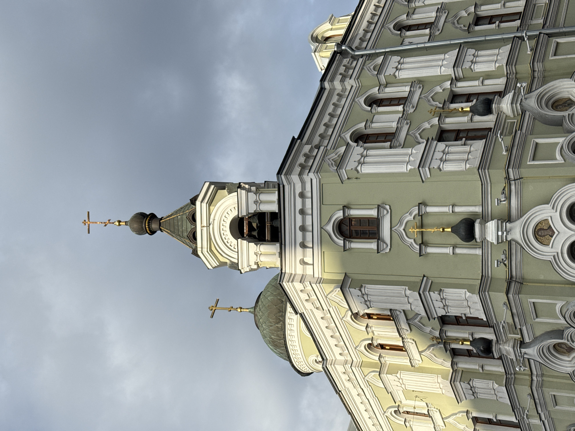The image shows the exterior of a pale green church with white architectural details, featuring multiple domes topped with crosses. The architecture is ornate with arched windows and decorative moldings. The sky above is overcast with dramatic clouds, and sunlight casts shadows on the building. The church has several cupolas, one larger green dome and a bell tower with bells visible. Smaller black domes with gold crosses are also present on the structure. The view is from a low angle looking up at the building. The location is identified as the Church of the Dormition of the Mother of God on Pokrovka street in Moscow, Russia. There are no people visible in the image.